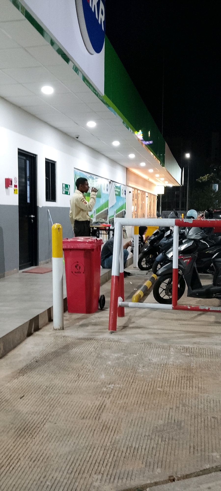 The image shows a man in a tan uniform drinking from a cup. He is standing near a red trash bin with a yellow pole in front of it. Behind him is a building with a green and white awning. There are several motorcycles parked behind a red and white barrier. The ground in the foreground is a textured concrete. The scene appears to be an outdoor area, possibly a gas station or convenience store in Gunung Putri, Indonesia, at night.