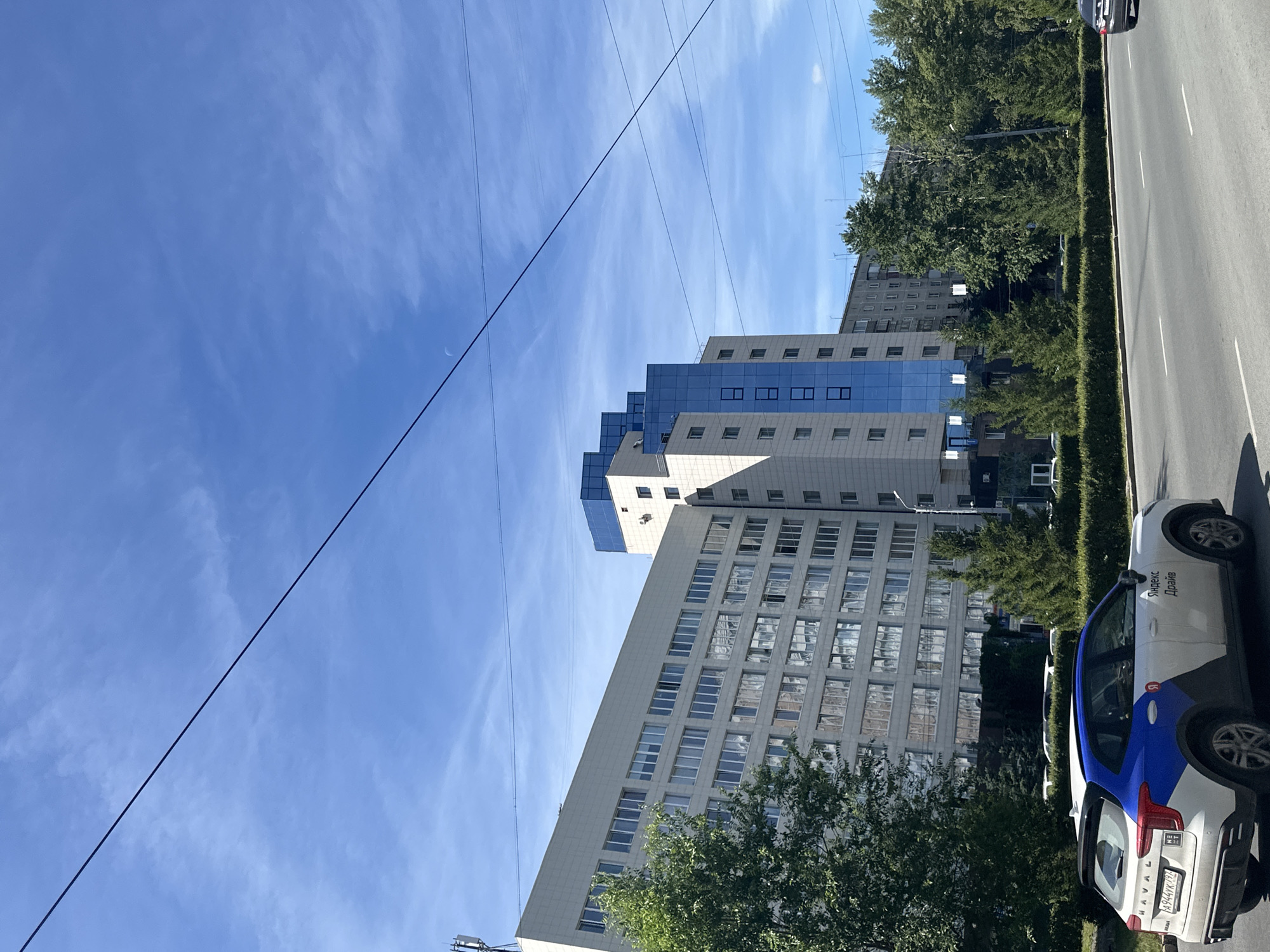 A wide shot captures a modern, light-colored building with a blue glass section on the right, towering over a tree-lined street. A white car with blue accents, possibly a service vehicle with logos, is parked on the right side of the road, facing left. The sky above is a clear blue with scattered white clouds and a faint crescent moon visible. The building features numerous windows, and the street appears to be a multi-lane road with a clear separation from the sidewalk and greenery. The overall scene depicts a daytime urban environment.