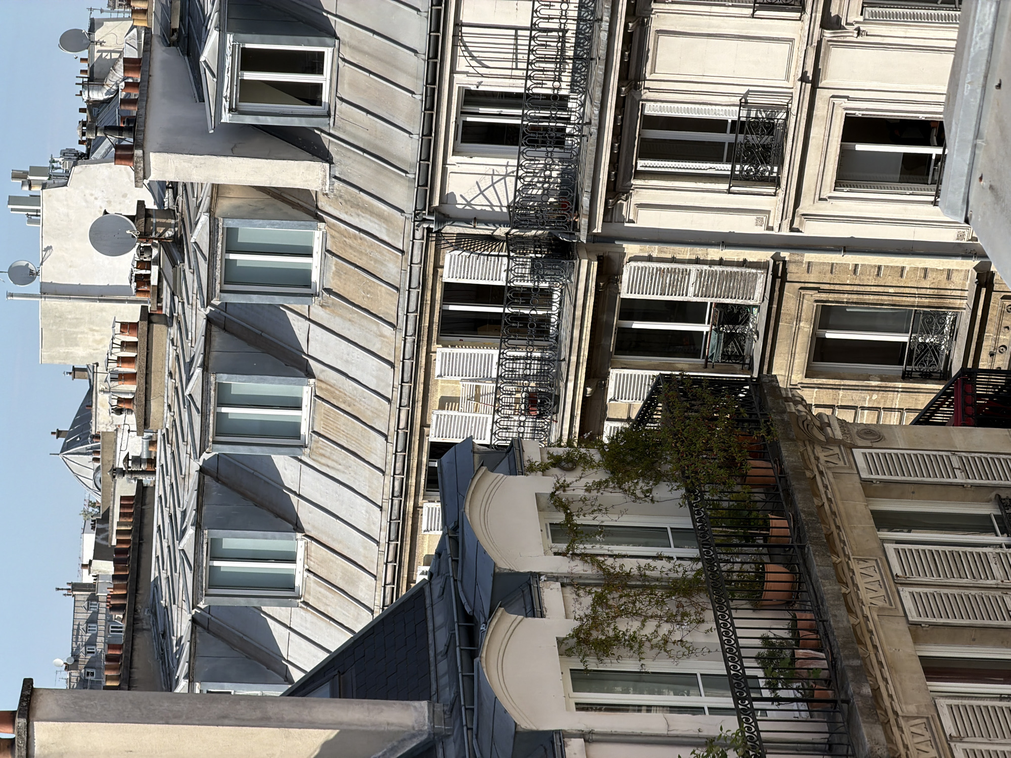 The image captures a high-angle, slightly skewed perspective of typical Parisian buildings and rooftops under a bright, clear sky. The scene is dominated by multiple layers of architecture, characteristic of the city.
In the foreground and extending across the middle, sloping zinc or metal rooftops with distinct ribbing patterns are prominent. Numerous brick chimney stacks of varying heights punctuate these roofs, and several dormer windows with white frames are visible, some open, others closed. White satellite dishes are mounted on various rooftop structures.
Below the rooftops, the building facades are visible, constructed from light-colored stone or painted stucco. They feature multiple windows, many equipped with traditional white louvered shutters, some of which are closed or partially open. Decorative wrought-iron balconies and window guards are a recurring element. On the right side of the image, a long, black wrought-iron structure, possibly a fire escape or an extended balcony, is adorned with green vines and terracotta pots. Architectural details, such as ornate carvings and moldings, are visible on some of the building fronts.
The strong sunlight creates sharp, diagonal shadows across the roofs and facades, indicating a clear day, likely late morning or early afternoon. No people or specific activities are visible, presenting a serene and architectural cityscape. There is no visible text in the image.