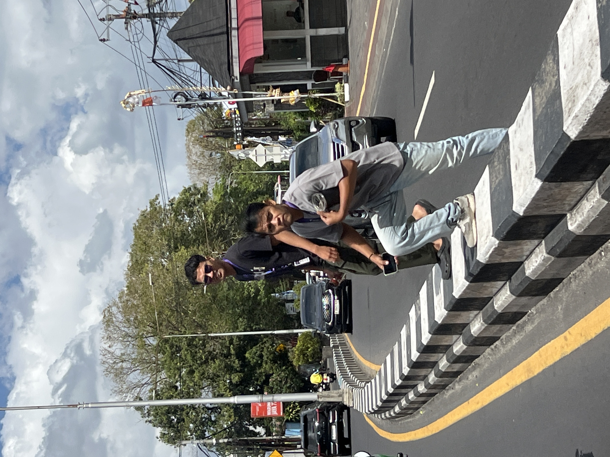This image depicts a street scene in Denpasar, Indonesia, likely near a busy intersection or road. Two men are the primary subjects. The man in the foreground, on the right, is leaning against a raised concrete barrier painted with black and white stripes. He is wearing a gray t-shirt and light blue jeans, with white sneakers. He is holding a cylindrical object, possibly a rolled-up poster or mat, and appears to be looking towards the viewer. The second man, standing further back and to the left, is wearing a black t-shirt and sunglasses. He is looking in the same direction as the first man, towards the road.
The environment shows a paved road with a yellow dividing line and a curb. Several vehicles are visible on the road, including black SUVs and a motorcycle with a rider in a yellow shirt. In the background, there are trees, utility poles with wires, and a small building with a red awning, which might be a kiosk or small shop. The sky is cloudy, indicating daytime. The scene suggests a casual street setting with some traffic and pedestrians.