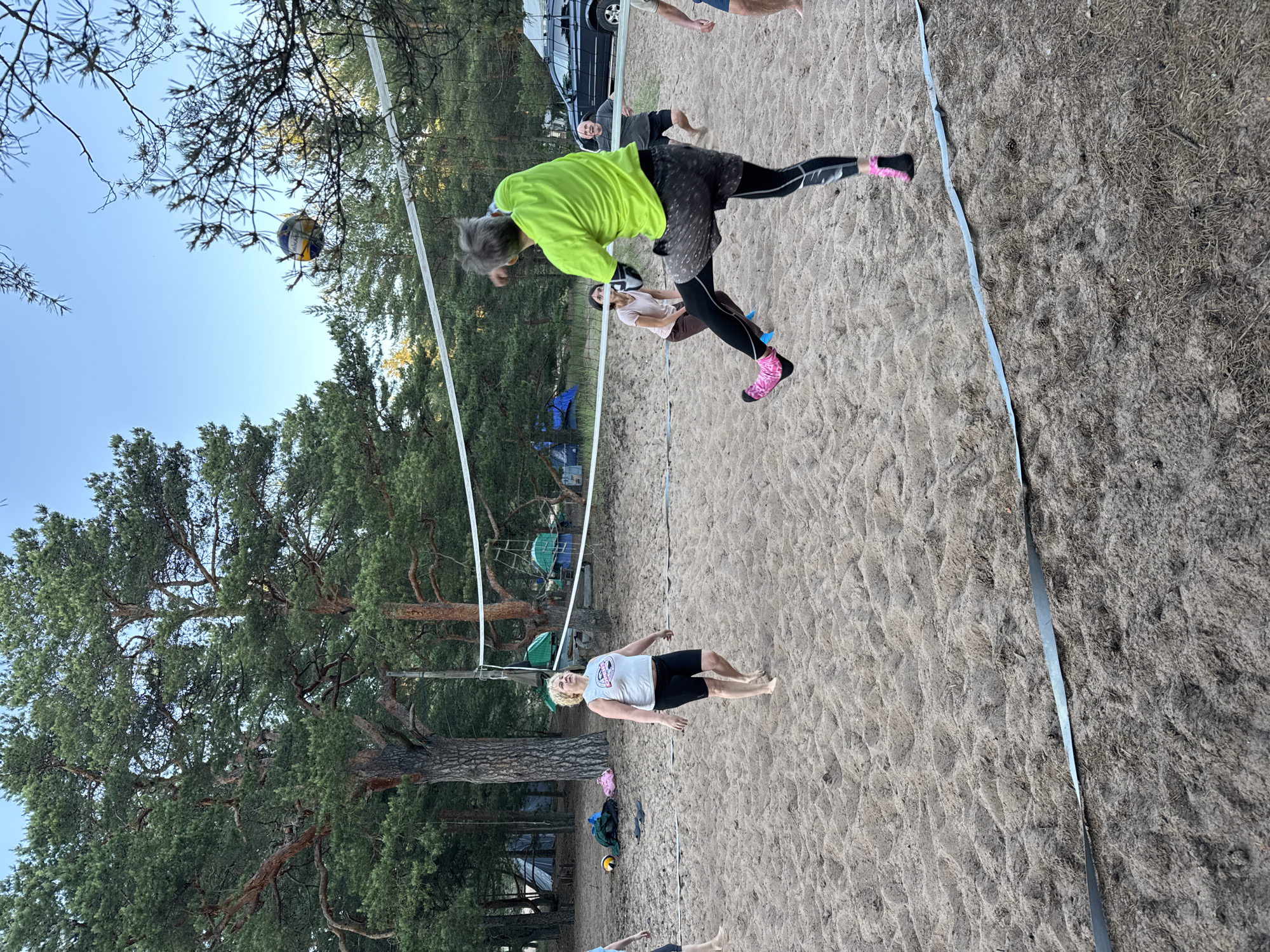In Ozerki (Krasnodolinskaya), Russia, a group of people are playing volleyball on a sandy court surrounded by pine trees. A volleyball net is set up, and a volleyball is in the air. One person, wearing a bright green shirt and patterned shorts, is mid-jump, appearing to hit the ball. Another person, wearing a white tank top and black shorts, stands on the sand. In the background, tents and a vehicle are visible, suggesting a campsite. The scene is set outdoors during daylight hours, with a clear sky.