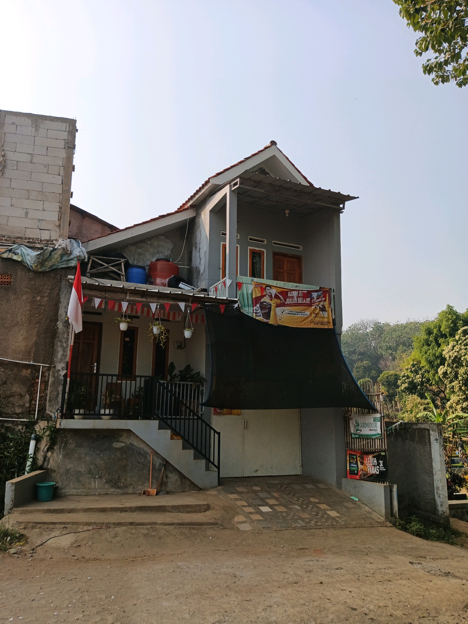 The image depicts the exterior of a two-story building, possibly a residence or a small commercial establishment, situated in a moderately dense urban or semi-urban environment. The building's facade is a mix of unfinished and painted concrete, with a prominent grey exterior paint on the upper levels. A red-tiled roof caps the structure, and a pitched roof extension is visible on the upper floor, supported by grey columns.
The ground floor features a large, white garage door or entrance, and a ramp leads up to it. Above the garage, a black mesh tarp is stretched across, creating a shaded area. Above this tarp, on the second floor, a banner with Indonesian text and images is displayed. A balcony area on the second floor is visible, with railings and windows.
To the left of the main building, an unfinished concrete block structure is present, suggesting ongoing construction or renovation. A red and white Indonesian flag is flying from a pole next to this structure. Small triangular red and white flags are strung along the balcony's railing. Hanging plants in white pots are also suspended from the balcony.
On the ground level, stairs with black metal railings lead up to the second floor. A teal bucket is placed on a concrete step to the left of the stairs, and a broom and dustpan are leaning against the wall beneath the stairs.
In the background, lush green trees and foliage are visible, indicating the presence of natural vegetation, possibly a hillside or a park. The sky is clear and bright, suggesting daylight. There are no people visible in the image. The overall scene suggests a residential area with some commercial activity or display. The presence of the Indonesian flag and the banners suggests a possible celebration or promotional event. The general impression is one of a functional, lived-in space with elements of local culture and economic activity.