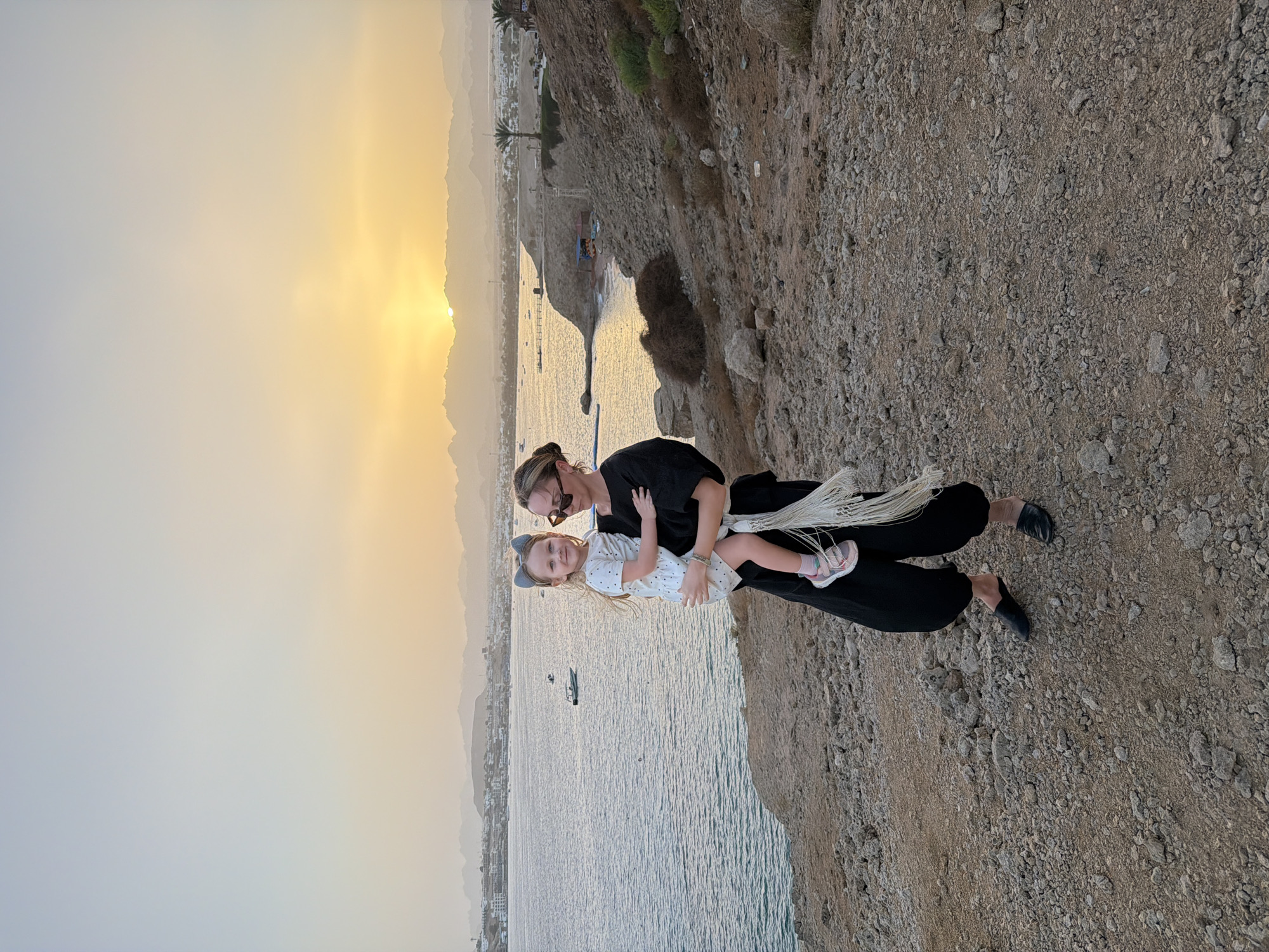 The image depicts a heartwarming scene at sunset in Sharm El Sheikh, Egypt, featuring an adult woman holding a young girl on a rocky cliff overlooking a calm body of water.
The woman has light-colored hair pulled back, wearing sunglasses on her head, a dark long-sleeved top, and dark pants with black flat shoes. A white, fringed accessory drapes over her arm. She holds a young girl with light-colored hair, dressed in a white polka-dotted dress, a headband with cat ears, and pink and white sneakers. The girl looks towards the viewer with a slight smile, while the woman maintains a neutral, perhaps subtly smiling, expression.
They are standing on rugged, rocky ground, characteristic of a coastal cliff, with the vast sea or ocean stretching out beneath them. Several small boats are visible in the distance on the water's surface. The distant coastline features buildings, a few palm trees, and hazy mountains receding into the horizon, indicative of the region's desert landscape.
The photograph captures the golden hour, with the sun positioned low on the horizon, casting a warm, radiant glow across the sky and reflecting on the water. The sky transitions from a soft blue on the left to vibrant golden and orange hues around the sun. The weather appears clear and serene. The activity captured is a posed moment, with the woman and child enjoying the scenic overlook during what appears to be a beautiful sunset. No visible text is present in the image.