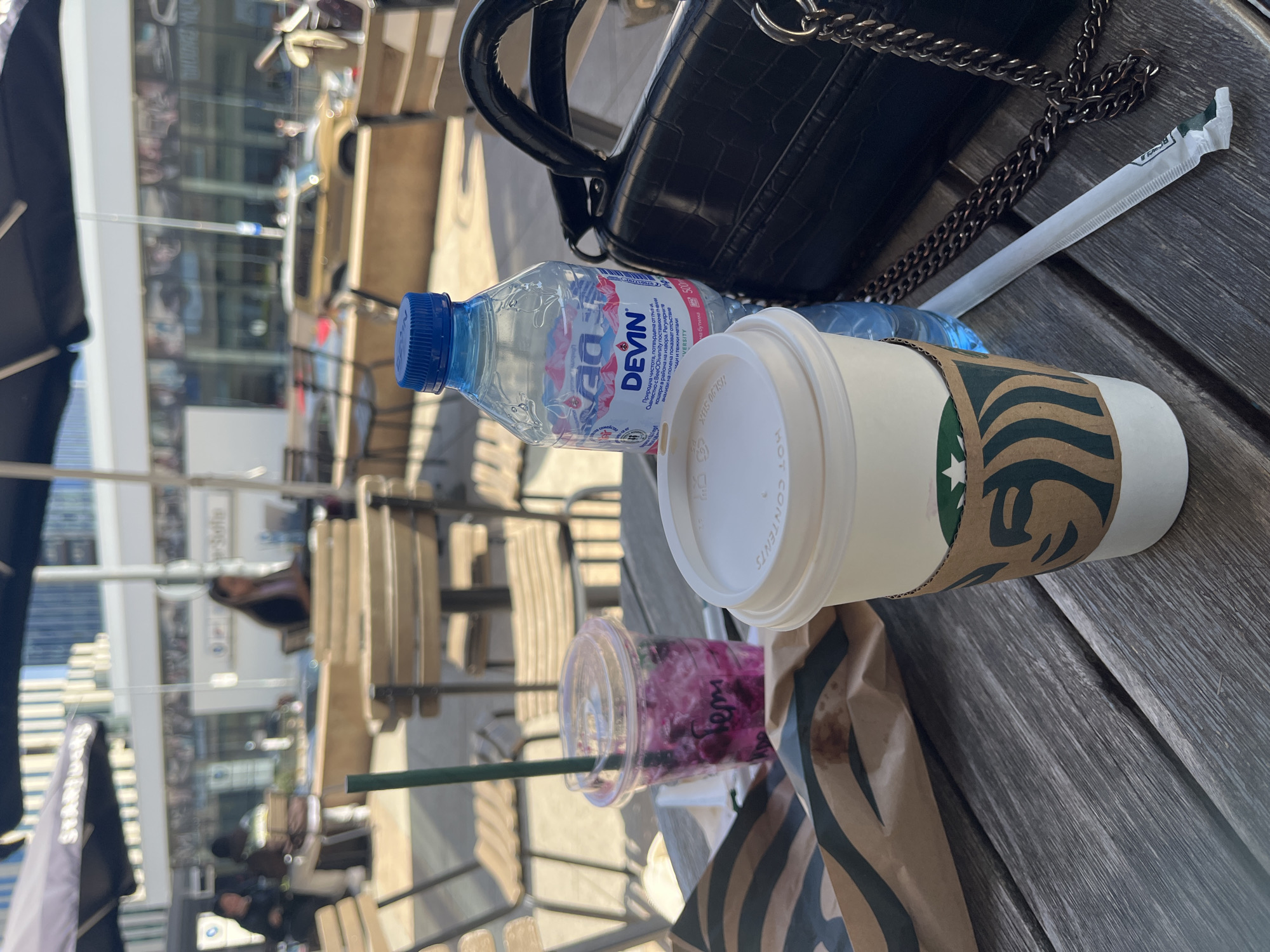 The image captures an outdoor cafe setting in Sofia, Bulgaria, during what appears to be a bright, sunny day. The scene is viewed from an elevated perspective, looking down at a wooden slatted table laden with drinks and personal items.
In the foreground, a black, textured handbag with a chain strap is placed on the table. Beside it, a plastic bottle of "DEVIN" brand mineral water with a blue cap is clearly visible. The bottle features Bulgarian text, partially legible as "Природна чистота" (Natural purity) and mentioning "BioDiversity" and "500" (likely milliliters). Next to the water bottle is a white Starbucks hot cup with a brown protective sleeve, displaying the iconic green Starbucks logo. The white lid of the cup has text, including "HOT CONTENTS" and "XO3-06-7SH". Further on the table, a clear plastic cup contains a vibrant purple-pink beverage, possibly a berry-flavored drink, with a green straw. A brown paper bag with the Starbucks logo is also partially visible, stained with a dark spill. A white paper-wrapped straw, partially opened, lies on the table near the bag.
The background is blurred, showing an urban environment with modern buildings, likely reflecting sunlight off their glass facades. Out-of-focus figures of people and outdoor cafe chairs with slatted wooden backs can be discerned. On the far left, a black banner or umbrella, partially visible, displays the "STARBUCKS" name, reinforcing the location as a cafe. The presence of the Bulgarian "DEVIN" water brand, combined with the Starbucks items and urban architecture, aligns with the specified location of Sofia, Bulgaria. The overall impression is one of a relaxed break in a bustling city environment.