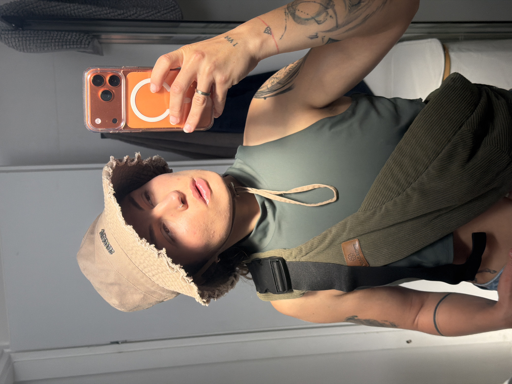 A person is taking a selfie with a phone in a room, possibly indoors, considering the presence of a wall and shelf. The person is wearing a beige bucket hat and a dark green tank top. The phone is in a clear case with a prominent orange circle design on the back, and the person has tattoos on their arms. A jacket of corduroy texture is draped over their shoulder. The presence of a wall and shelf behind the person suggests an interior location. There is no indication of the city of Kiryat Bialik, Israel.