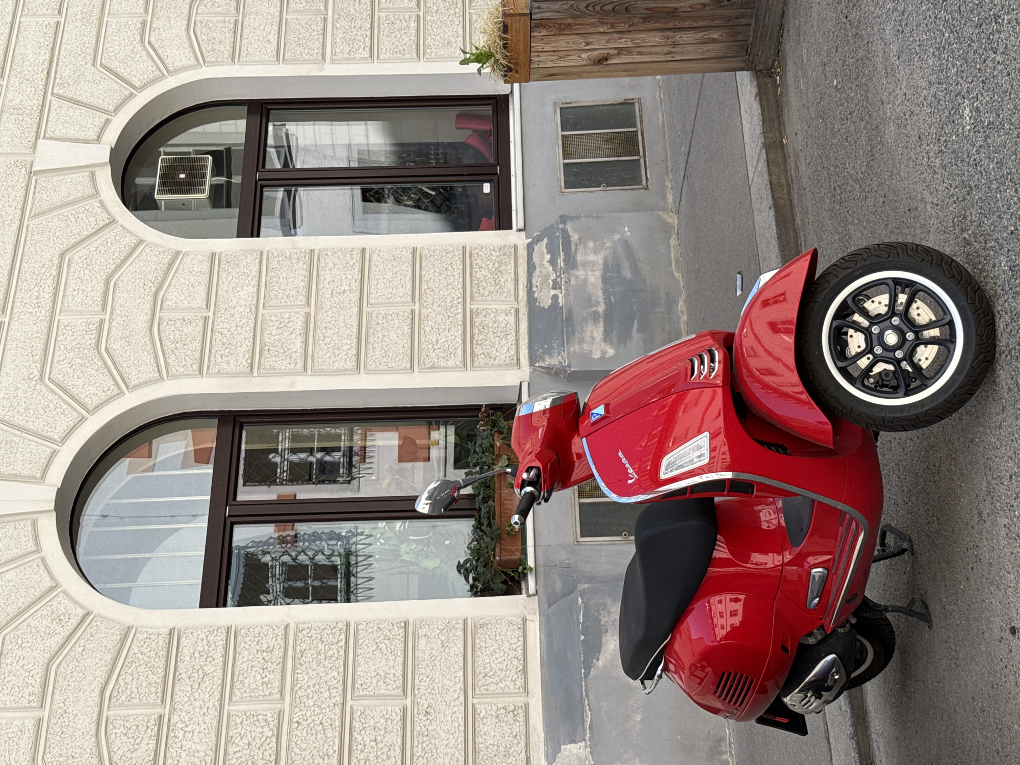 A bright red Vespa scooter is parked on a paved street in front of a light-colored building with arched windows. The building has a textured facade, resembling rough-hewn blocks. The windows are dark-framed, with the left window reflecting a building with grid-like patterns, and the right window showing what appears to be red shoes and a black object inside. The scooter is positioned at an angle, facing slightly to the left. It has a black seat, chrome accents, and a black wheel with white trim. A kickstand is down. To the right of the scooter, a wooden planter box with a small plant sits on the sidewalk. The environment suggests an urban setting, possibly a European city.