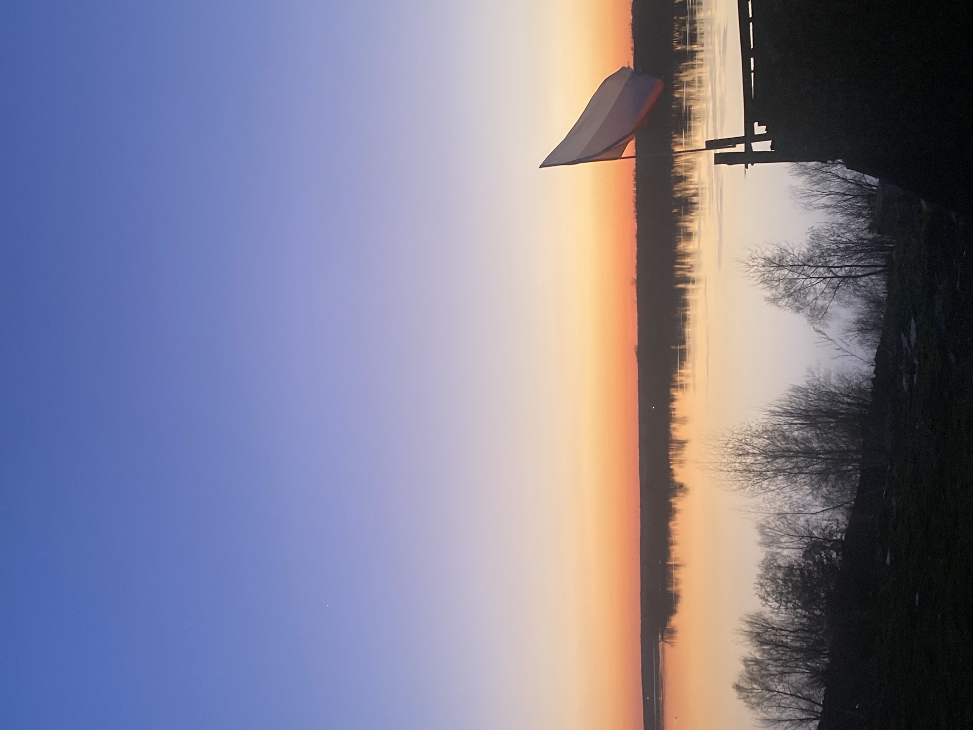 The image shows a sunset in Ольховка, Russia, reflected in a body of water. The sky transitions from blue to orange. A Russian flag is visible on what appears to be a pier. The silhouettes of bare trees and vegetation are on the right side of the image.