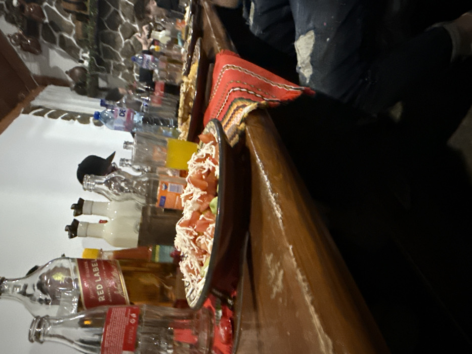 An indoor dining scene in Terziysko, Bulgaria, features a wooden table laden with food and beverages, suggesting a social gathering. In the immediate foreground, a dark ceramic bowl holds a salad composed of diced red tomatoes, green components, and shredded white cheese. To the left of the salad, an uncapped Johnnie Walker Red Label whisky bottle, partially full, stands next to an empty glass Coca-Cola bottle. Further back, several other empty clear glass bottles, a small orange condiment bottle, and a glass containing a yellow liquid are present. A woven textile with red, black, yellow, and green stripes serves as a table runner, partially draped over the table edge to the right. The wooden table surface exhibits signs of wear.
In the mid-ground, additional plates of food, various beverage bottles including a "DEVIN" branded water bottle, and multiple glasses are arranged along the table. Several individuals are partially visible and out of focus in the background, seated around the table. One person is identifiable by a black baseball cap worn backward. The lower right foreground shows the upper thigh of an individual wearing dark denim jeans with a noticeable tear or distressed patch. The background includes a white wall and a stone-clad wall adorned with pottery items, beneath a wooden ceiling beam.