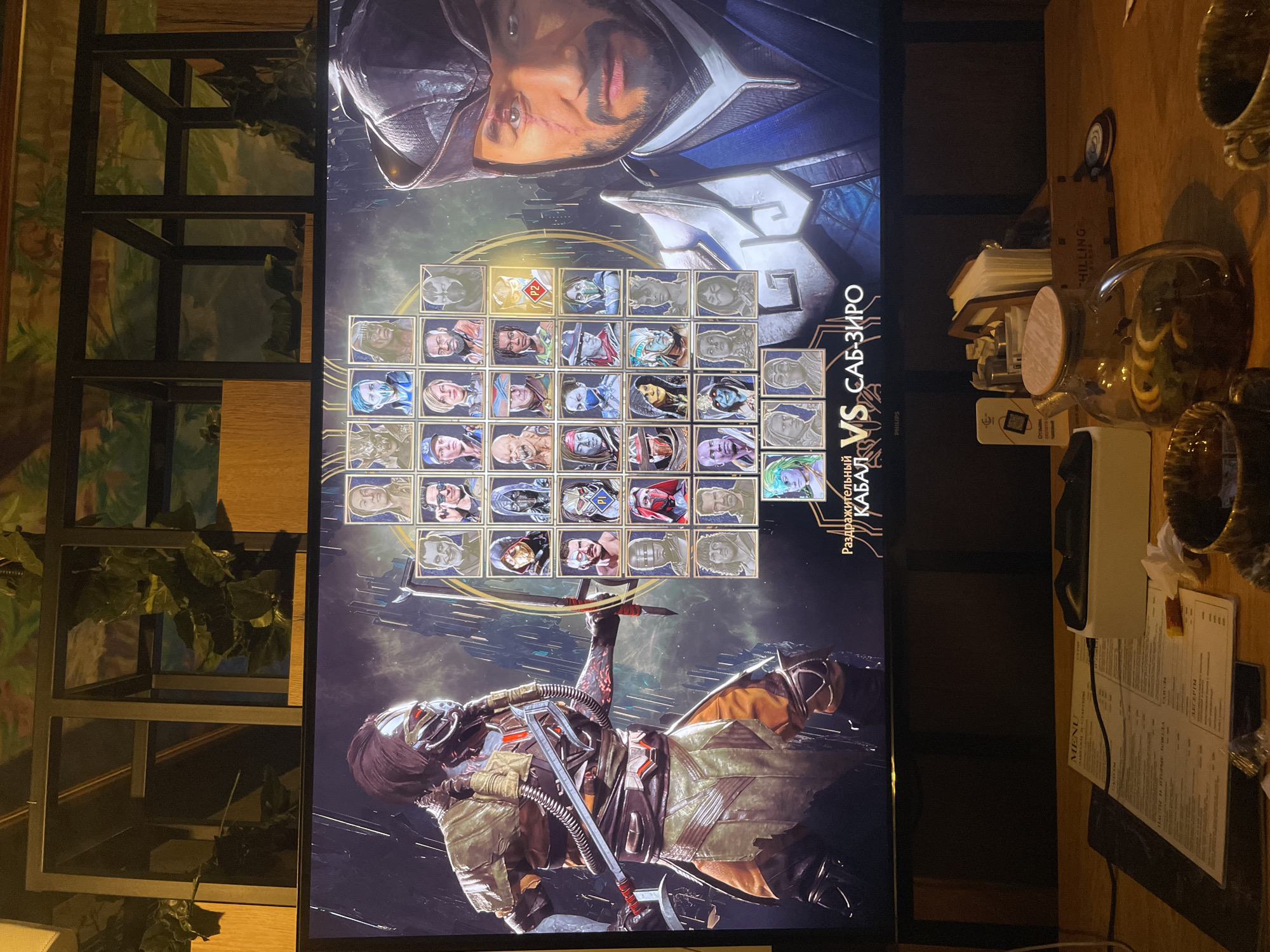The image depicts a Mortal Kombat 11 character selection screen, likely being played in a public space. The game shows "Kabal" versus "Sub-Zero" in Russian text. The physical environment includes a table with restaurant menus and a teapot, suggesting the setting is a cafe or restaurant. A metal shelf with plants is visible to the left of the screen. No people are visible in the shot. The location is indicated as Kazan, Russia by the Russian text in the image.