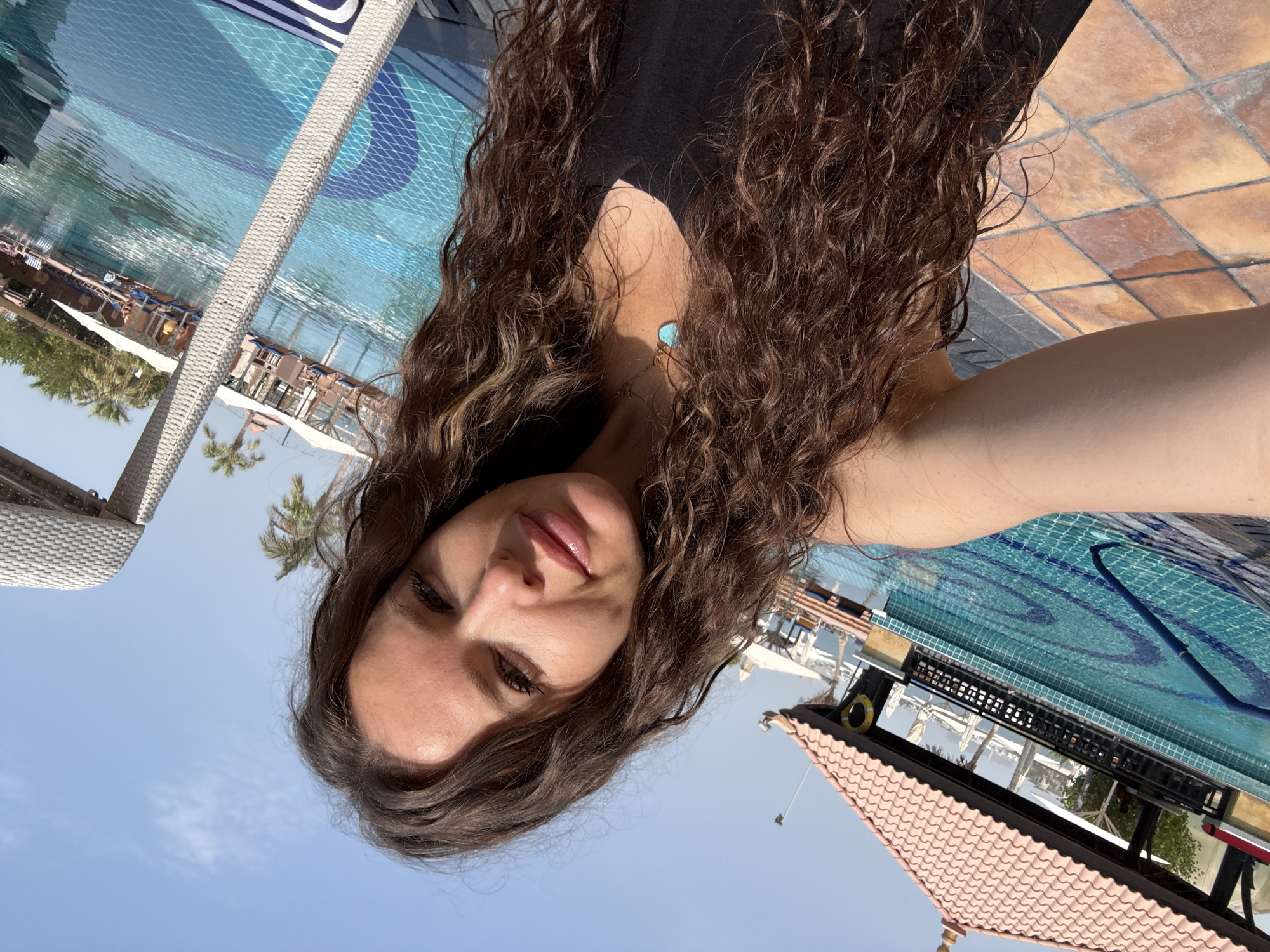 In Dubai, United Arab Emirates, a woman with long, curly brown hair takes a selfie by a swimming pool. She is wearing a black top and a turquoise pendant necklace. The pool has a tiled bottom with a mosaic pattern and is surrounded by a tiled deck and lounge chairs. In the background, there are palm trees, cabanas, and a building with a terracotta-tiled roof. The sky is clear and blue.