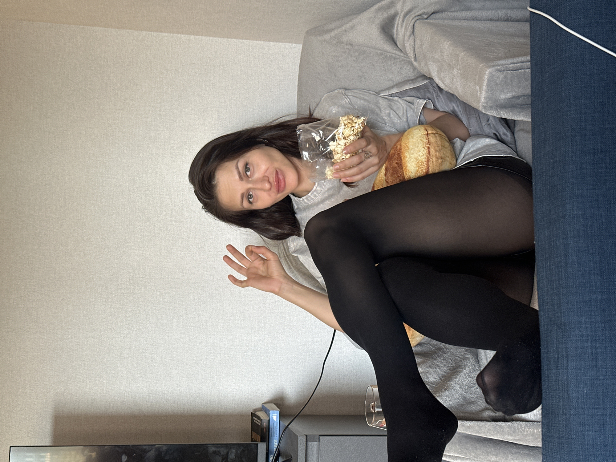 The image, presented rotated counter-clockwise, captures a young woman relaxing indoors on what appears to be a couch or bed. She has long, dark hair and is looking directly at the camera with a subtle smile or smirk. She wears a light grey t-shirt and black sheer pantyhose or tights.
Her left hand holds a clear plastic bag filled with light-colored snacks, likely popcorn or nuts. Her right hand forms an "OK" gesture. Next to her legs is a large, plush object resembling a loaf of bread or a baguette. She is seated on light grey upholstered furniture, partially covered by a matching grey blanket. To the right, a section of blue fabric furniture is visible, over which a white cable is draped. A black power cord runs from behind her right hand downwards.
In the bottom left of the frame, below her legs, a dark electronic device (possibly a media console) with a reflective surface is visible, alongside a few books. A blue book spine clearly shows the text "BCE". A clear glass or cup rests on the surface near her right leg.
The background features a plain, textured light-beige or off-white wall. The lighting suggests a bright, indoor daytime setting. The woman appears relaxed, content, and playful, posing for the photograph in a domestic environment. This scene is set in Murino, Russia.