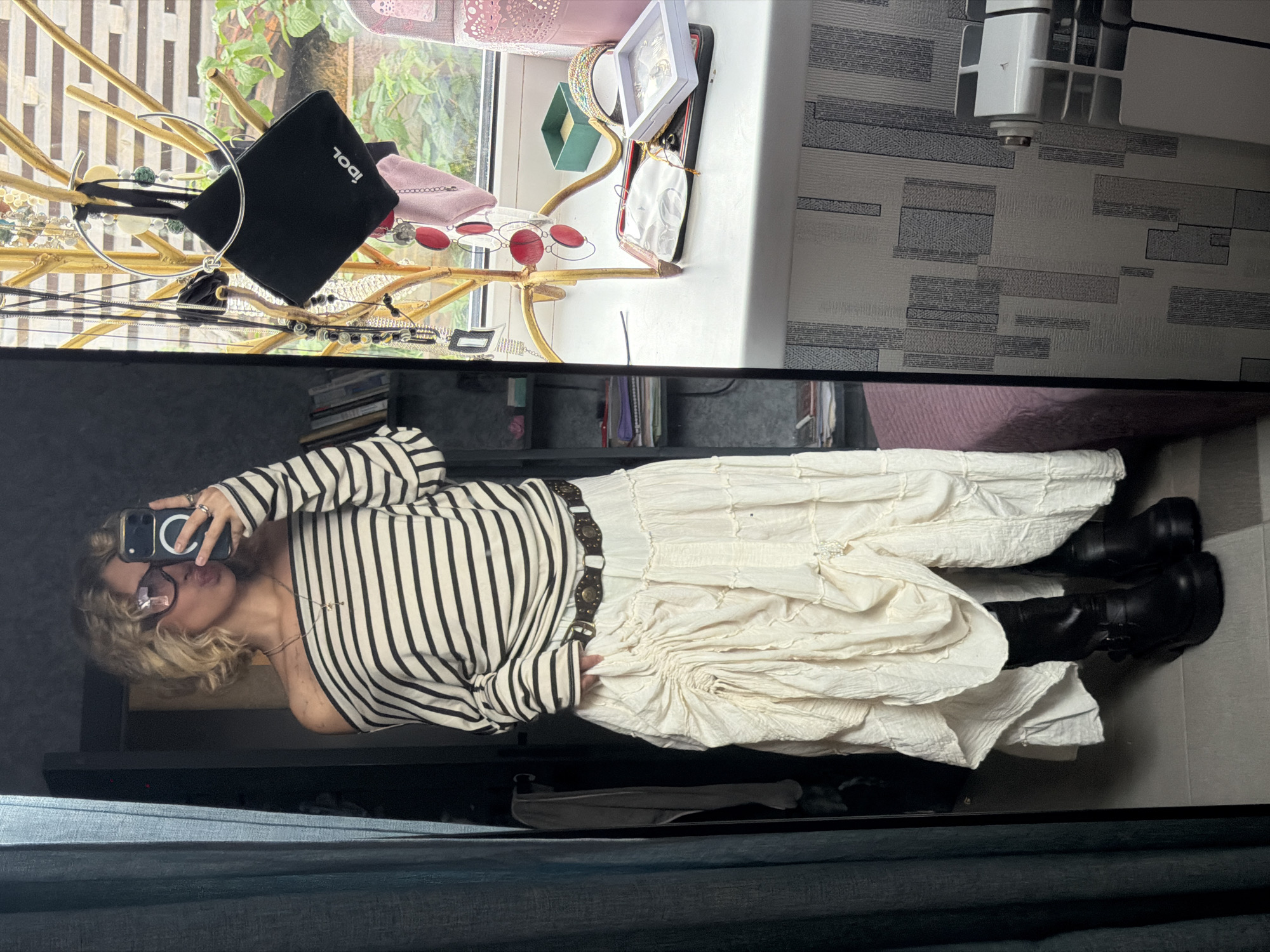 A young woman stands in front of a mirror, taking a selfie. She has blonde, curly hair and is wearing large, square-framed glasses. She is wearing an off-the-shoulder striped long-sleeved top, a cream-colored, tiered maxi skirt with ruching and a decorative belt, and black platform boots. The background shows a window with greenery outside, a jewelry stand with various necklaces and a small black bag, and a dresser or shelf with decorative items. To the left, a teal curtain is visible. The floor appears to be tiled. The image is taken indoors, likely in a residential setting, possibly a bedroom or dressing area. No specific landmarks or indications of the city of Kamenka, Russia, are present in the image.