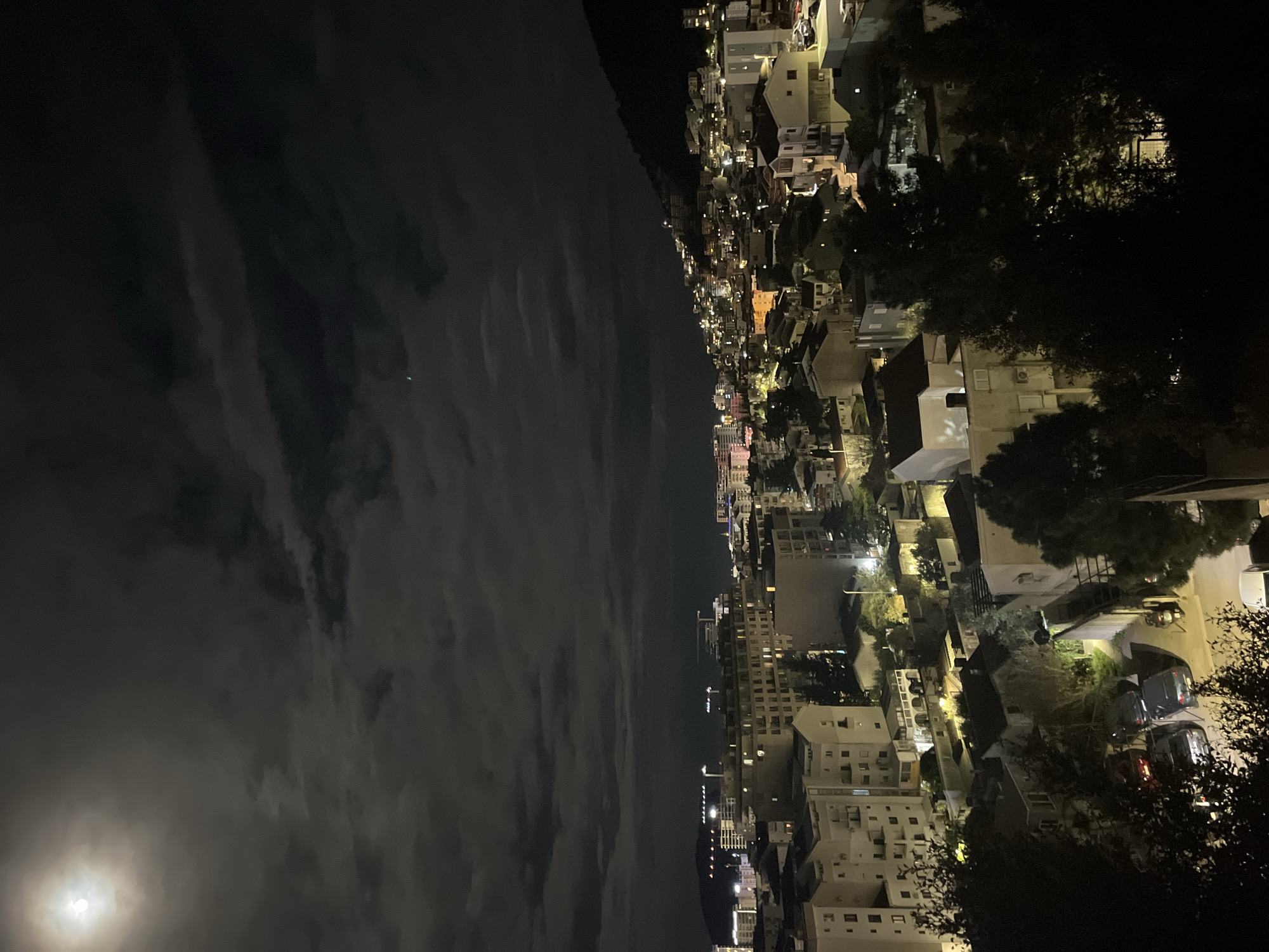 The image depicts a nighttime view over the city of Budva, Montenegro. A dark, cloudy sky with a visible moon dominates the left side of the frame. On the right, the city sprawls across the landscape, with buildings ranging in height and architectural style illuminated by artificial lights. Some trees are visible among the buildings on the right. The scene is generally dark, with the city lights providing the primary source of illumination.
