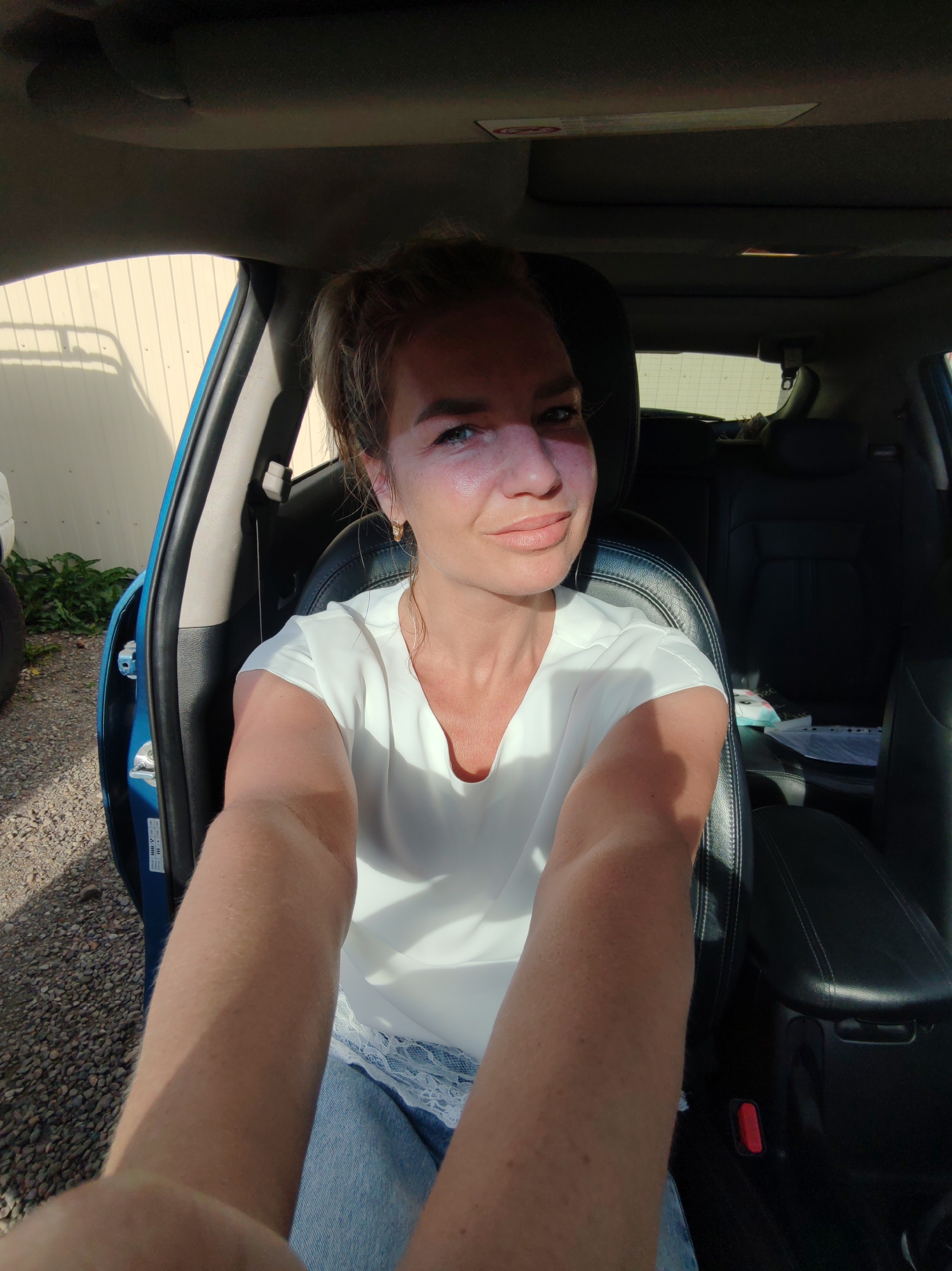 A woman is sitting in the driver's seat of a car, with the car door open on the driver's side. The woman is blonde and is wearing a white shirt and blue jeans. She is holding a phone in her right hand, as if taking a selfie. The car's interior is visible, including black leather seats and a center console. The exterior of the car is blue. Outside the car, there is a gravel driveway and a light-colored building with horizontal siding. The image appears to have been taken in Paratunka, Russia, likely during daylight hours, as evidenced by the lighting and shadows.