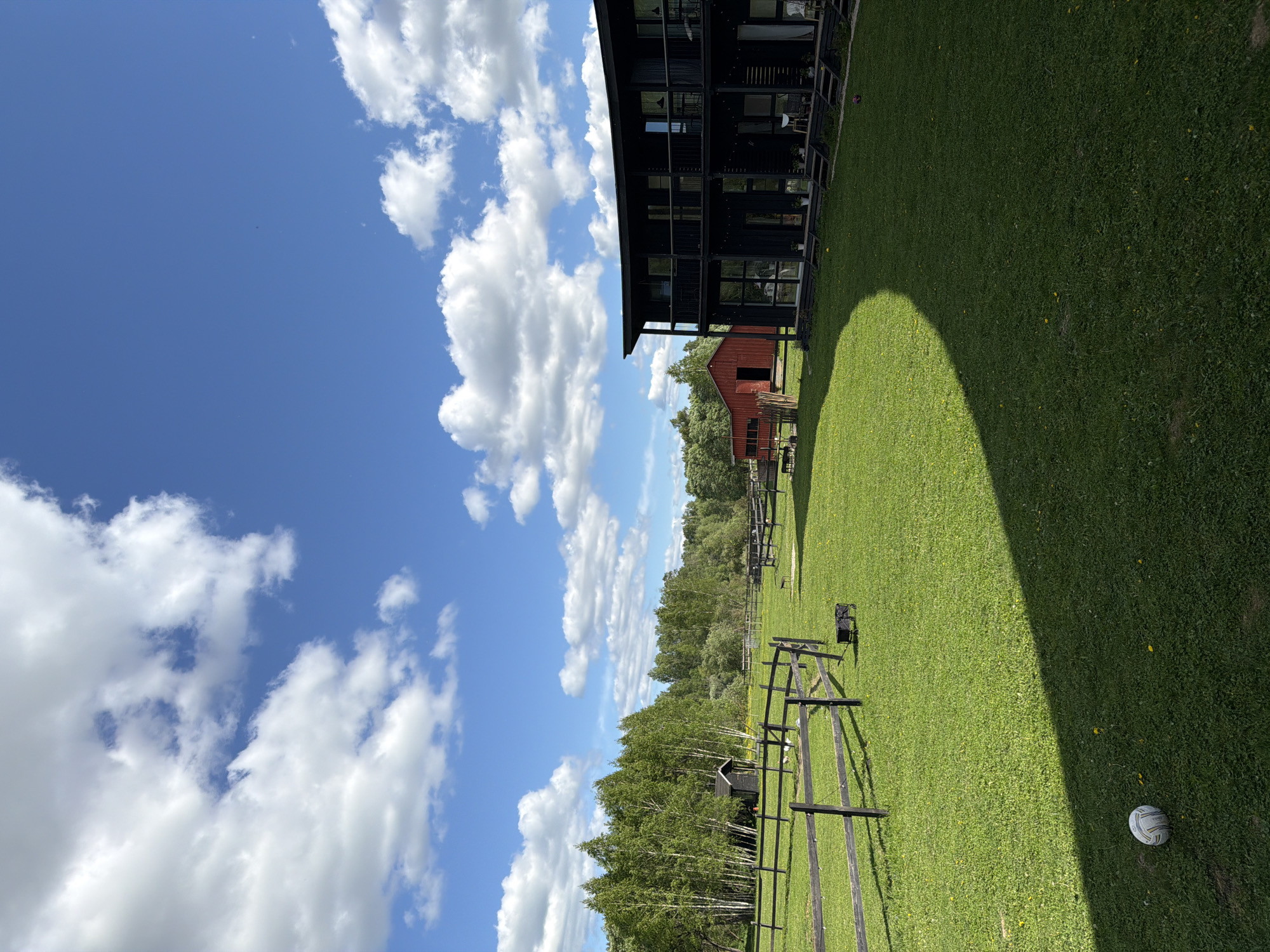 The image presents a rural scene in Prudki, Russia. A large, modern black building stands adjacent to a well-maintained grassy field. A smaller red barn is visible in the background, nestled among trees. A rustic, black wooden fence runs across the field. In the distance, a dense line of trees separates the field from the horizon. The sky is bright blue with scattered white clouds. A shadow from the black building falls across the grass. A soccer ball rests on the grass in the foreground.