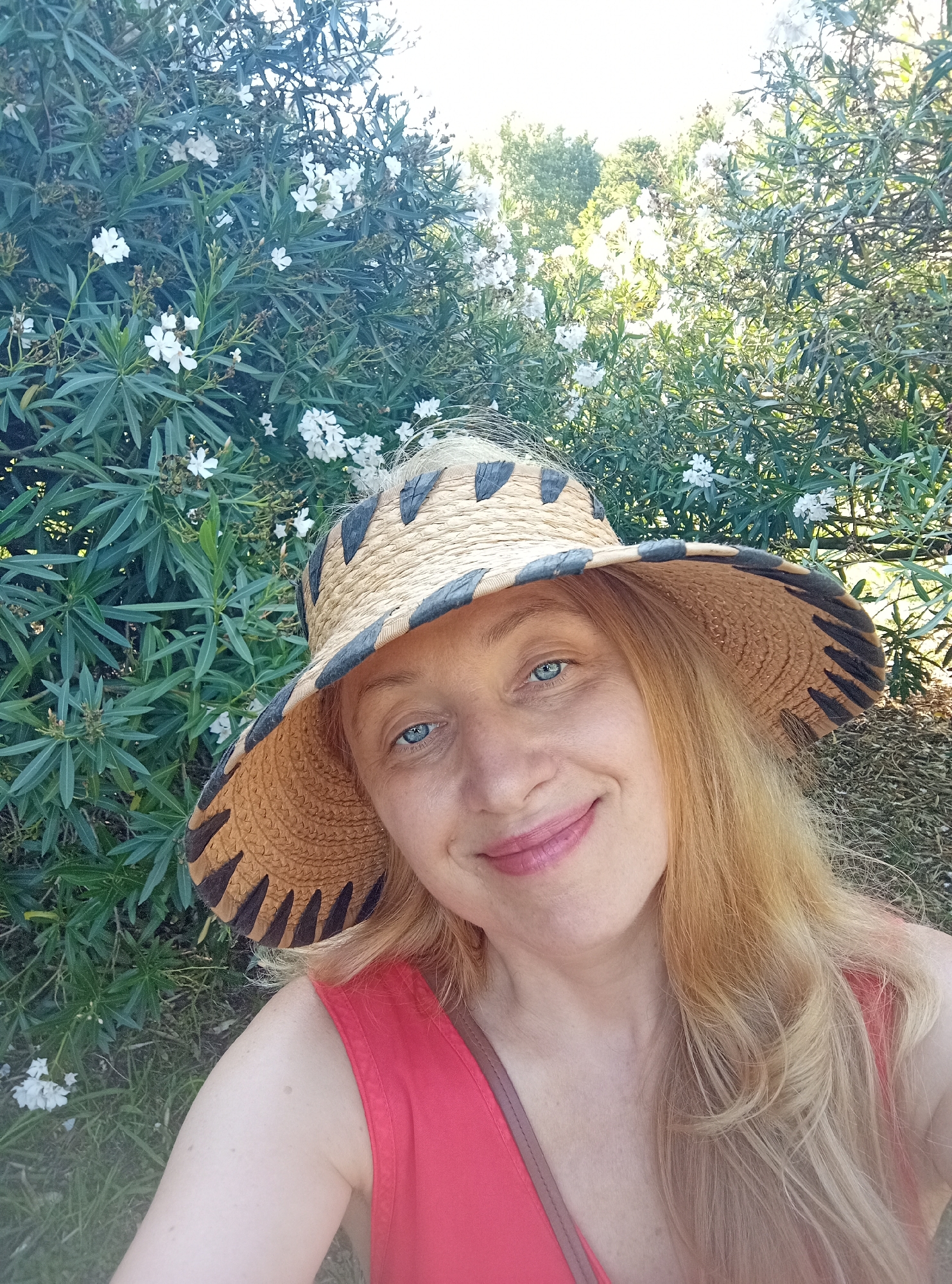 In a sunlit outdoor setting, a woman with blonde hair and striking blue eyes smiles at the camera, wearing a wide-brimmed straw hat and a coral-colored top. She is positioned in front of a dense green bush dotted with small white flowers, suggesting a garden or park environment, potentially in Lisbon, Portugal, given the warm, Mediterranean-like foliage. The lighting is bright, casting soft shadows and highlighting the textures of the hat and the woman's skin. The background consists of more greenery and dappled sunlight filtering through leaves, creating a serene and natural atmosphere.