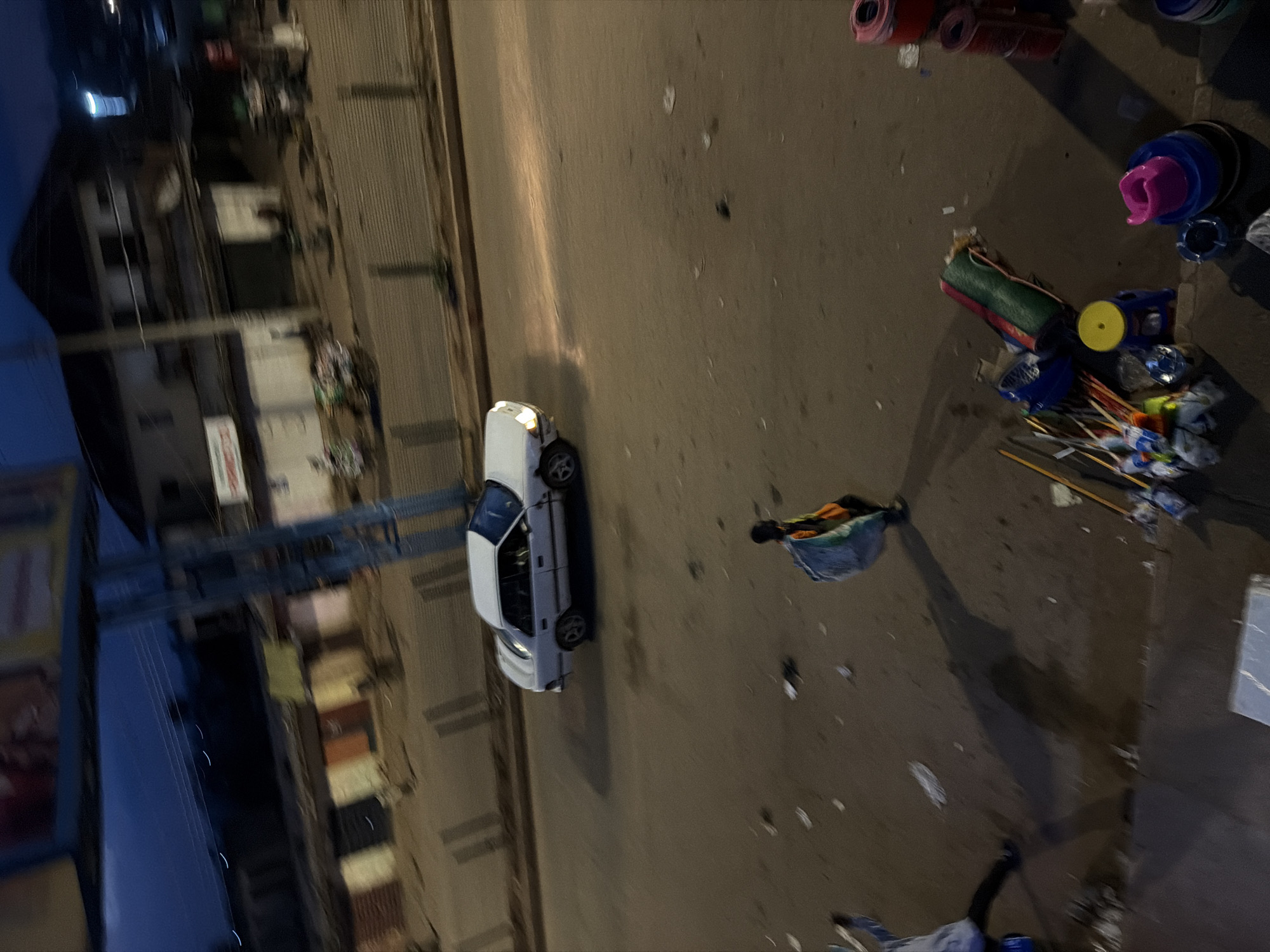The image depicts a street scene in Bukuru, Nigeria, during twilight. A white sedan with its headlights on is driving on the asphalt road. In the foreground, a person is walking away from the camera, carrying a large, light-colored bag over their shoulder. Another person is partially visible to the left, also appearing to be walking. The roadside is lined with shops and buildings, some with visible signage. Various objects, including buckets, mats, and brooms, are scattered on the ground near the roadside. The ground appears damp in some areas, suggesting recent rain. The overall lighting suggests the end of the day.
