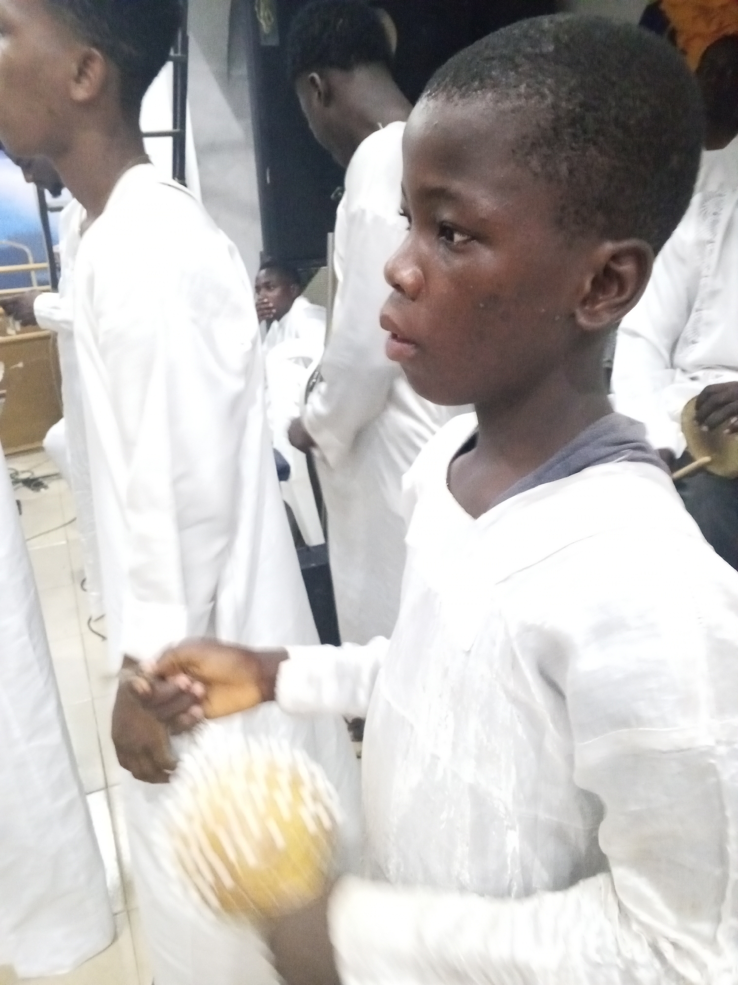 In Lokoja, Nigeria, a group of individuals, including a young boy, are gathered indoors. All are wearing white garments, suggesting a ceremonial or religious event. The boy, positioned in the foreground, holds a maraca. Other individuals are visible in the background, some seated. The interior setting appears simple, with light walls and a few pieces of furniture.