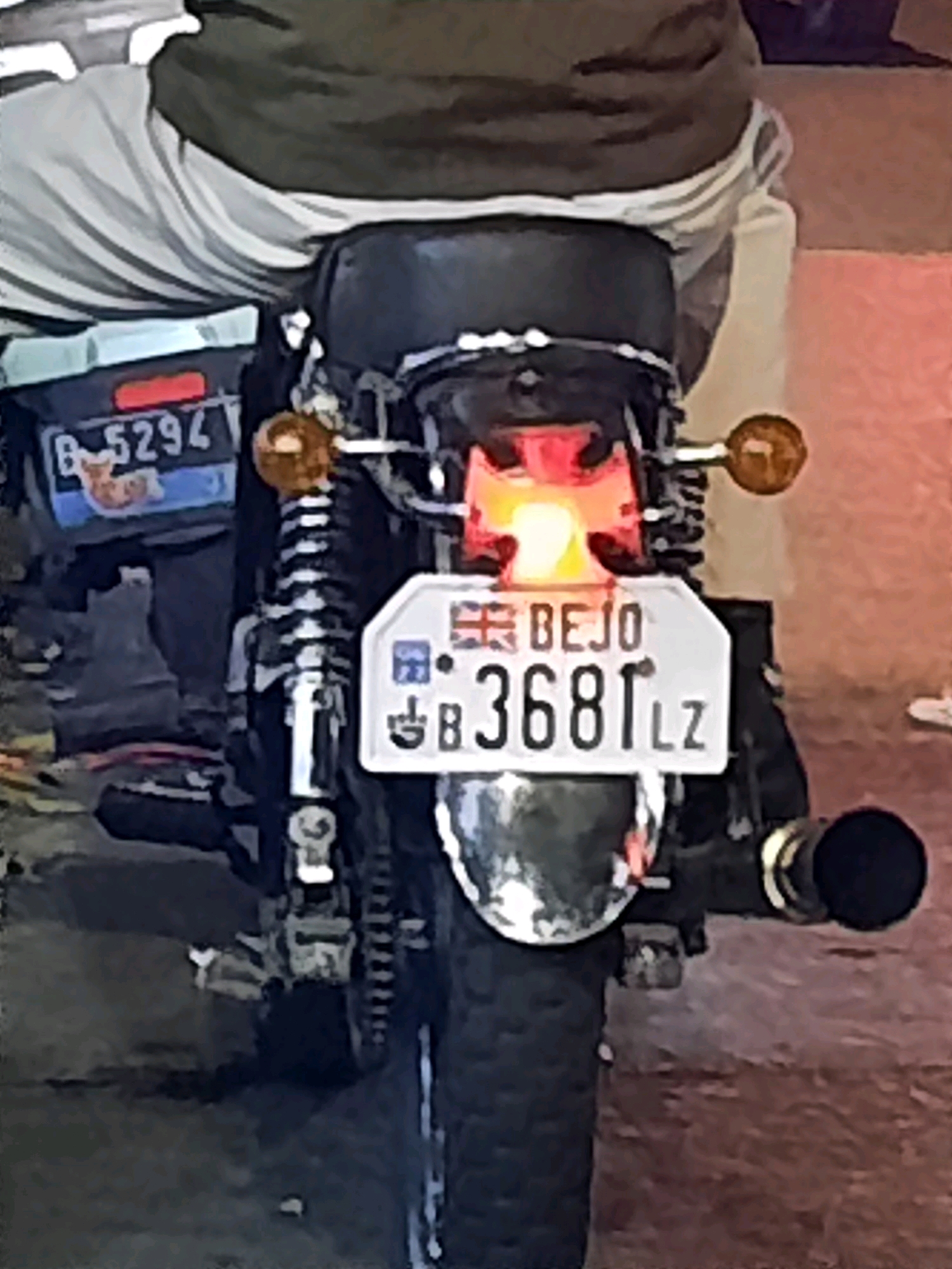 The image shows the rear view of a motorcycle parked outdoors. The motorcycle has a license plate that reads "BEJO 3681 LZ". There is also another license plate visible on the side of the motorcycle that reads "B 5294". The rider is partially visible from the back, wearing a dark jacket over light-colored pants. The background appears to be a street or parking area. The overall scene is dimly lit. This image was taken in East Jakarta, Indonesia.