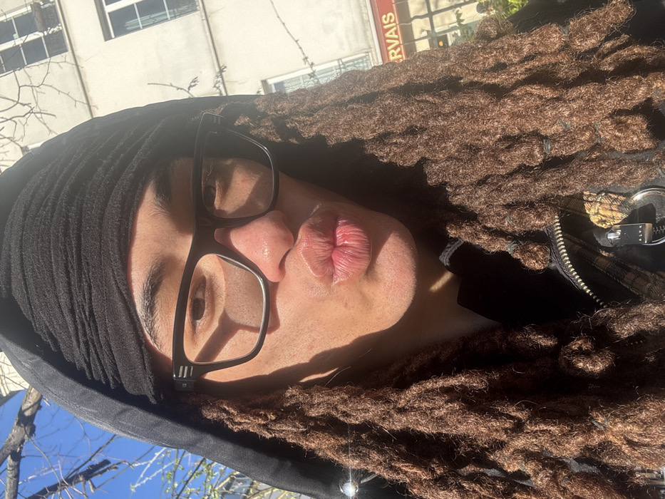 The image displays a close-up outdoor portrait of an individual in Paris, France. The person has medium-dark skin, long brown dreadlocks partially secured with black bands, and wears black-framed glasses featuring three horizontal lines on the temples. They are dressed in a black hooded garment with a black fabric wrapped around their head beneath the hood. The individual is making a "kissy face" with pursed lips, facing directly towards the camera. A portion of a dark jacket with a checkered pattern and a metallic zipper pull branded "ALYX" is visible on their chest. Direct sunlight illuminates the scene, creating defined shadows on the person's face. In the background, a light-colored, multi-story building typical of Parisian architecture is visible, featuring several windows. A sign on the building's lower level clearly displays "ST GERVAIS" in red letters on a light background, identifying the location as the Saint-Gervais neighborhood. Bare tree branches are discernible against a clear blue sky.