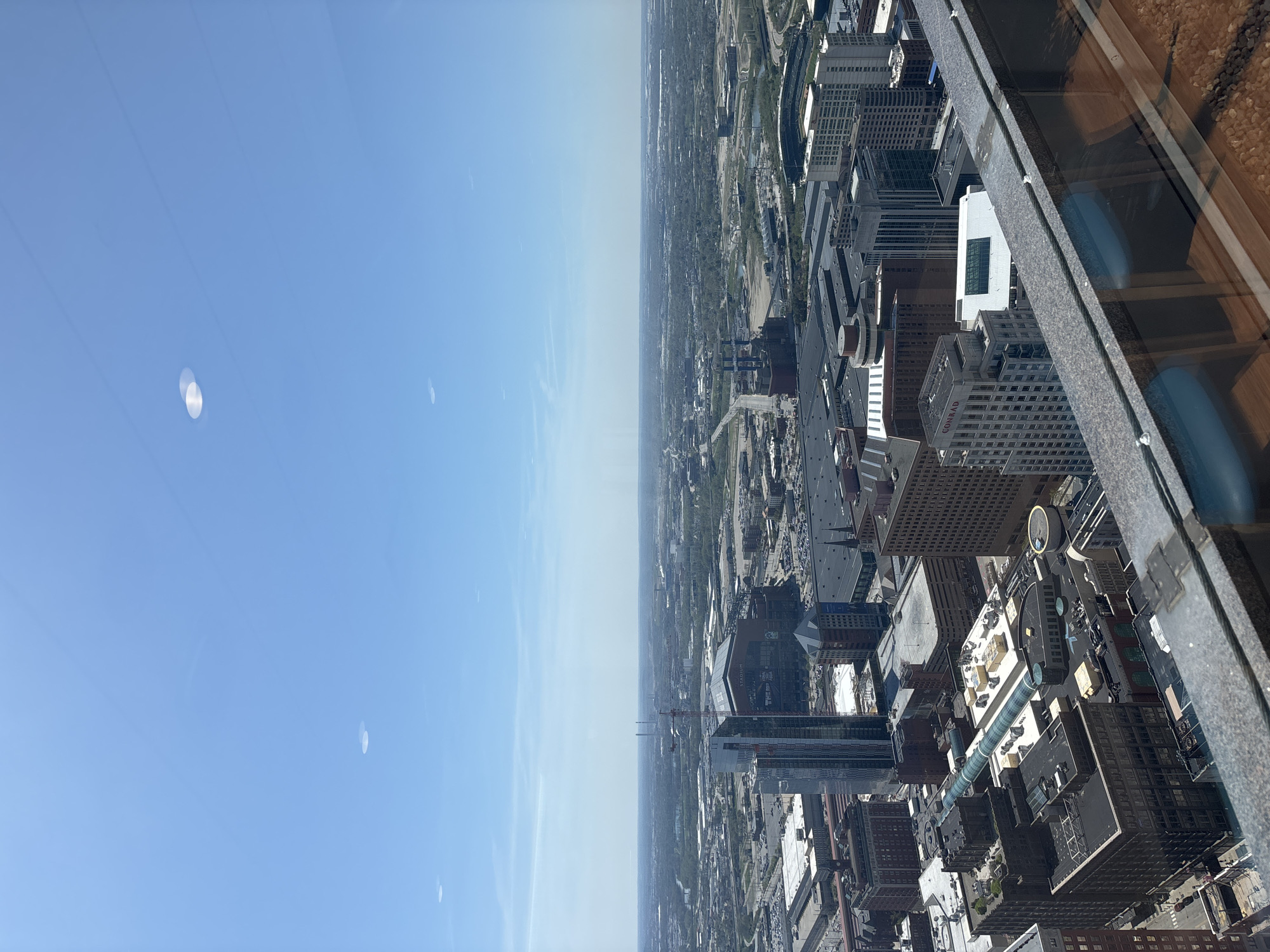 Here is a detailed description of the image:
The image presents an aerial view of Indianapolis, Indiana, taken from a high vantage point, likely within a skyscraper. The scene captures a clear, bright sky on one side and the city landscape on the other. The skyline features a dense concentration of high-rise buildings and skyscrapers, typical of a major urban center.
Buildings: The architecture varies from modern glass structures to older brick buildings. A prominent white building with a large rectangular window near the top can be seen, along with other buildings that have varying textures and heights.
Roads: Multiple roads and streets are visible, running through the urban grid, with what appears to be traffic on them.
Environment: The landscape includes both natural elements, like trees and green spaces, and human-made structures. The overall environment is urban, characterized by dense construction and a network of infrastructure.
Other Features: The view is framed by an element of the building from which the picture is taken, including a window frame and a partial view of an interior space (possibly a ledge), indicating the photo was shot from inside a building.
In summary, the image is a high-angle shot of Indianapolis, capturing the city's skyline, buildings, roads, and overall urban environment, likely taken from a tall skyscraper.