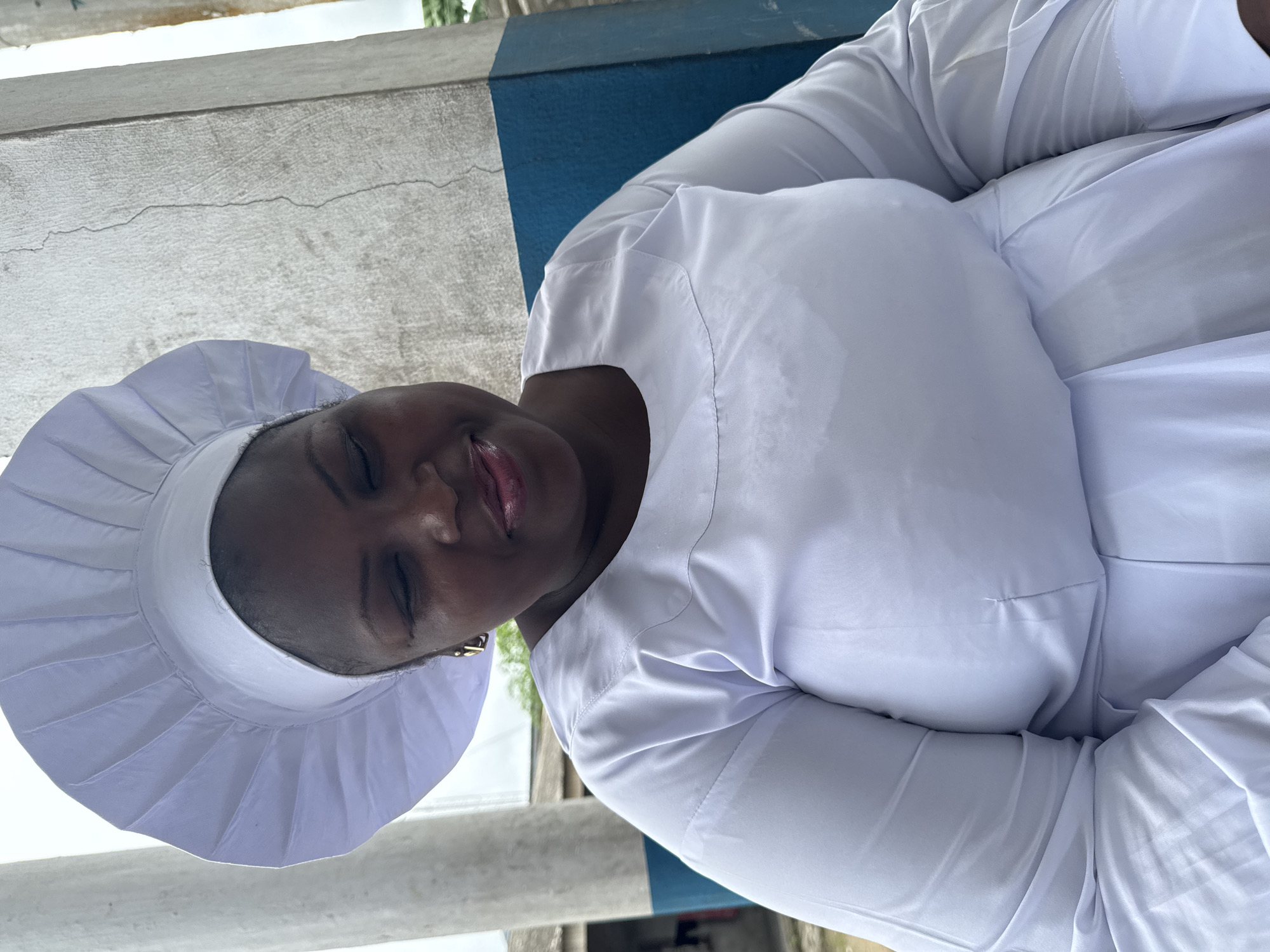 The image shows a medium shot of a woman wearing a white uniform and a white cap, likely a religious or ceremonial garment, indoors with a partially visible outdoor background. The woman is positioned in the center of the frame, facing slightly to the right, with her eyes closed and a gentle smile on her face. She has dark skin and is wearing gold hoop earrings. The uniform is loose-fitting and made of white fabric, with pleats visible on the cap and folds in the dress.
Behind the woman, a blue and white striped wall is visible. The blue stripe runs horizontally across the wall, with a rough, textured gray section above it. The lighting suggests an overcast day or an indoor setting with diffused light. In the background, to the left, there are glimpses of what appears to be buildings and vegetation, indicating an outdoor environment.
This scene is consistent with being located in Badagry, Nigeria, a city known for its historical significance and strong religious presence. The style of architecture and the typical weather conditions in the region align with the visual cues present in the image.