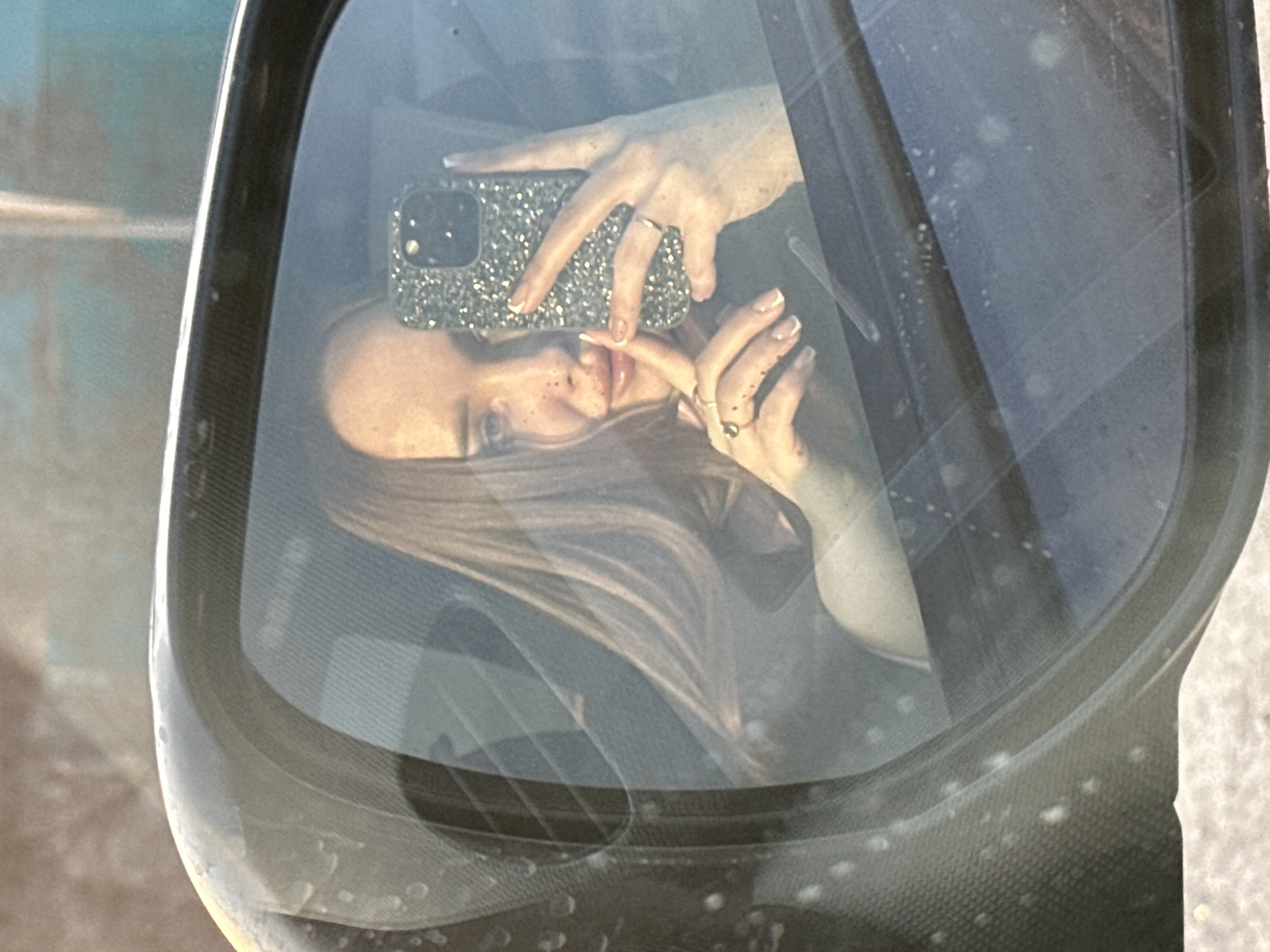 The scene, located in the city of Pirogovo, Russia, presents a frontal view of a female individual reflected in the driver's side mirror of a vehicle. The female has long, light brown hair and visible freckles on her face. Her lips appear to be covered with a gloss or lipstick. She is actively holding a smartphone, encased in a highly reflective, glitter-covered case, in her right hand, positioned as if taking a photograph of her reflection. Her left hand is partially visible, displaying a ring on the left ring finger and another on the left pinky finger, complementing a ring on her right ring finger. Her fingernails exhibit a French manicure style. The dark-colored mirror housing and surrounding frame show numerous small, light-colored specks, indicative of dust or dirt. The reflection also reveals portions of the vehicle's dark interior and an obscured outdoor background, with a light-colored ground surface visible at the bottom of the mirror.