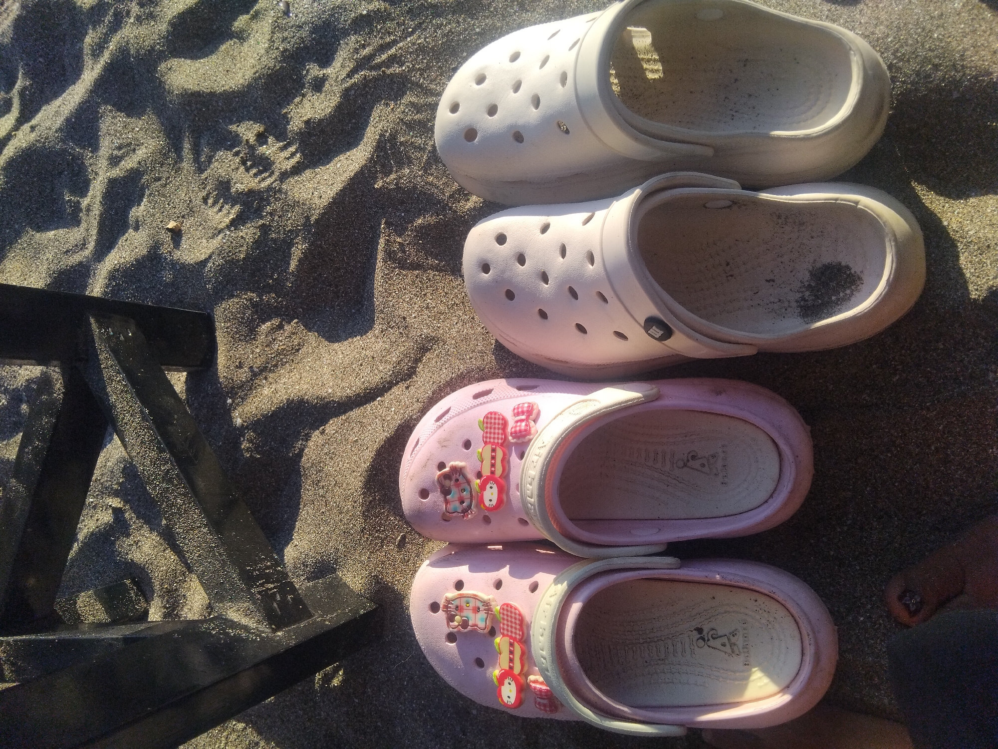 Four pairs of Crocs-style clogs are arranged on sandy ground, captured from an overhead perspective. The top two pairs are off-white or light grey and appear to be adult-sized. The bottom two pairs are pink and child-sized, adorned with various colorful charms.
The sandy surface, possibly a beach, shows distinct ripples and textures, indicating it's an outdoor setting. Strong sunlight casts clear, defined shadows, suggesting it's a sunny day, likely during the morning or late afternoon given the angle of the light.
The white clogs show some wear and accumulated dirt or sand inside, particularly a dark spot in one of them. The pink clogs feature several cartoon-like charms, including a brown cat-like figure, red fruits (possibly apples or strawberries), checkered bows, and a white bunny or cat face. Inside the pink clogs, the sole has a textured pattern with some embossed text or a logo, though it's largely unreadable. A small black logo, possibly an "M" or similar, is visible on the side pivot of one of the white clogs' straps.
In the bottom left, part of a black metal object, likely a leg of a chair or table, is visible, partially buried in the sand. On the far right, a bare foot with painted toenails (dark polish with a glittery accent) is slightly visible next to the pink clogs. The scene evokes a relaxed, leisure setting, typical of a beach outing.