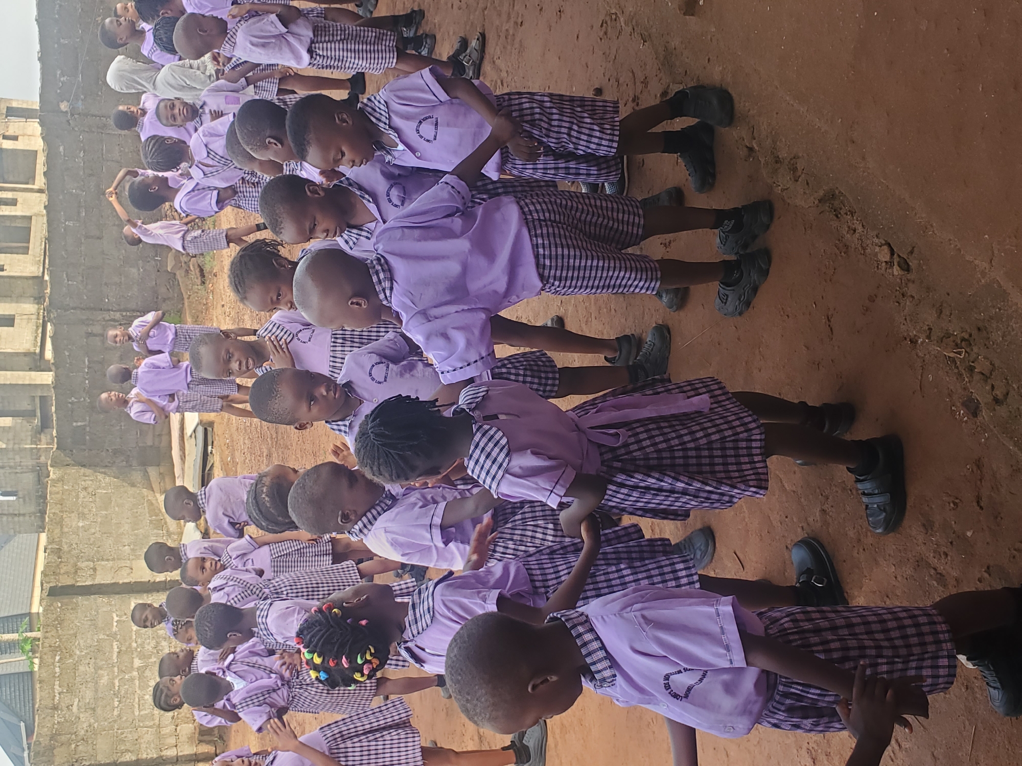 A group of children in school uniforms are lined up on a dirt surface next to a building. The children are wearing a lavender shirt with a black and white checkered skirt or shorts and black shoes. It is daytime. The location is Ukwu-Abwa, Nigeria.