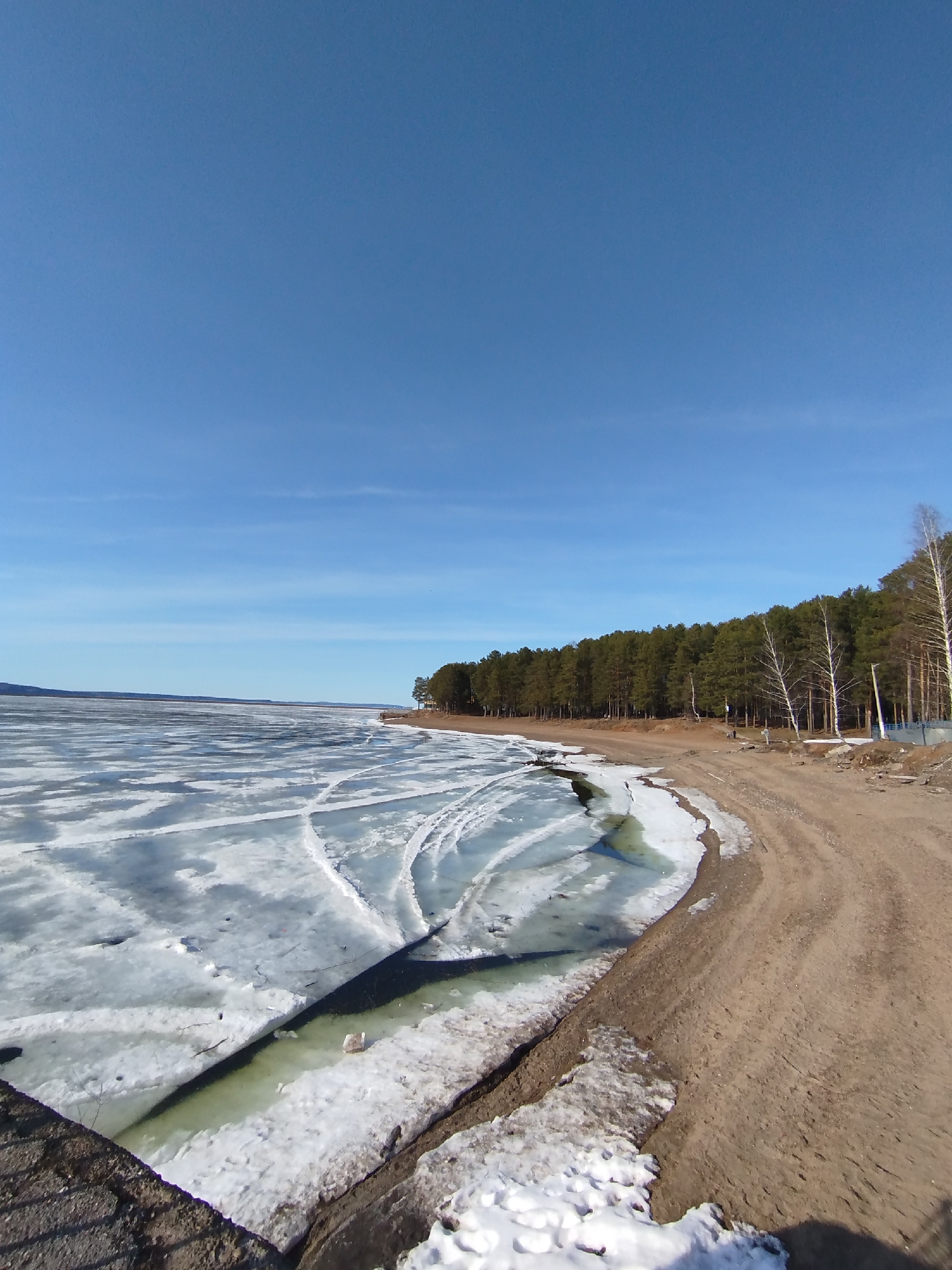 The image depicts a partially frozen body of water along a sandy shoreline in Chaikovsky, Russia. Large sheets of ice cover the water's surface, with visible cracks and open water areas. A sandy beach borders the water, transitioning to a forest of coniferous and deciduous trees. The sky is a clear blue. Patches of snow are visible on the ground near the shoreline.