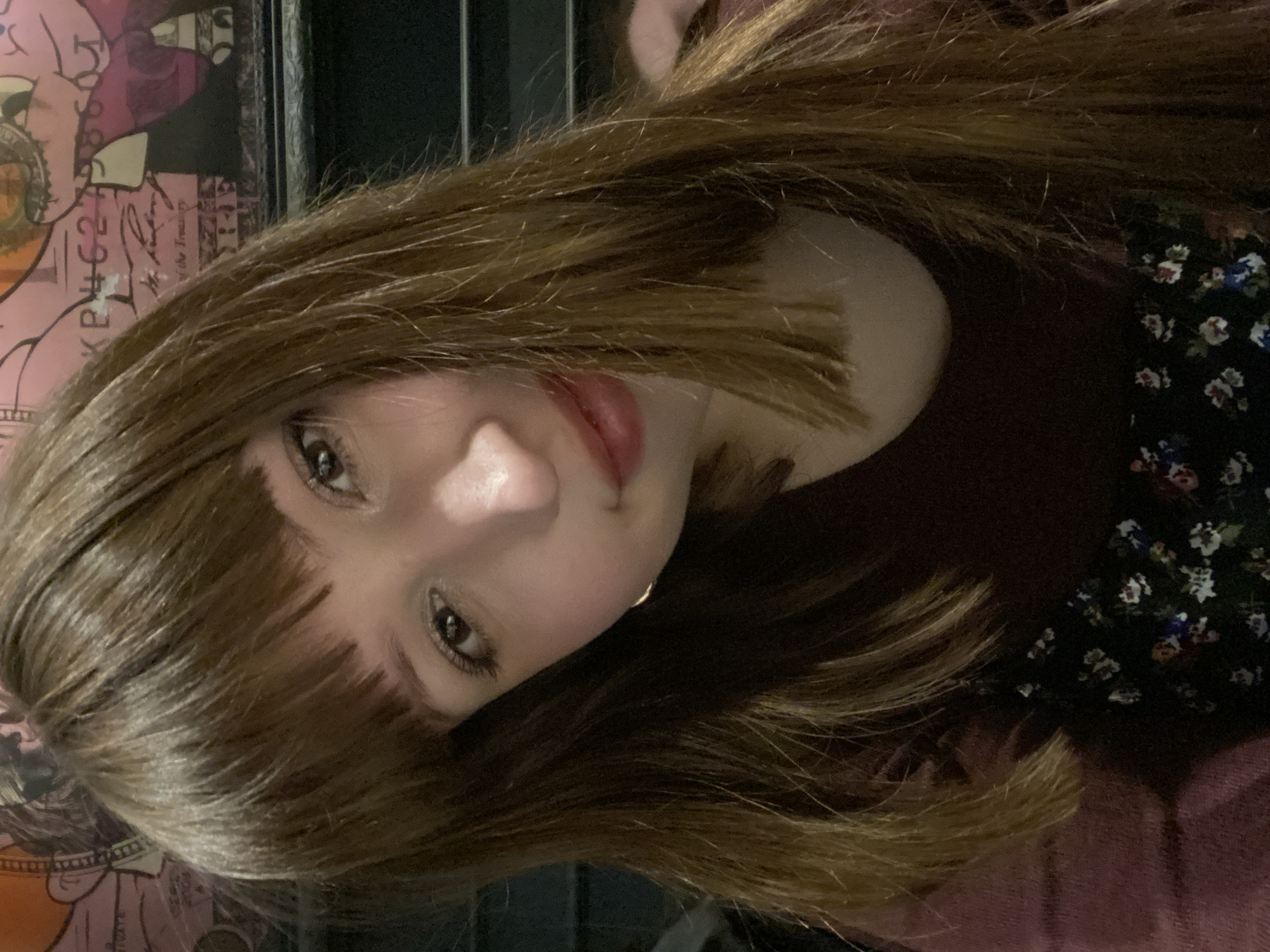 A close-up portrait shows a person with brown hair and bangs, light skin, and visible eyes and lips. The person is wearing a dark-colored top with a floral pattern. A map or poster with faded colors and illegible text is partially visible in the background. The location in the city of Chaikovsky, Russia, is not discernible from the image.