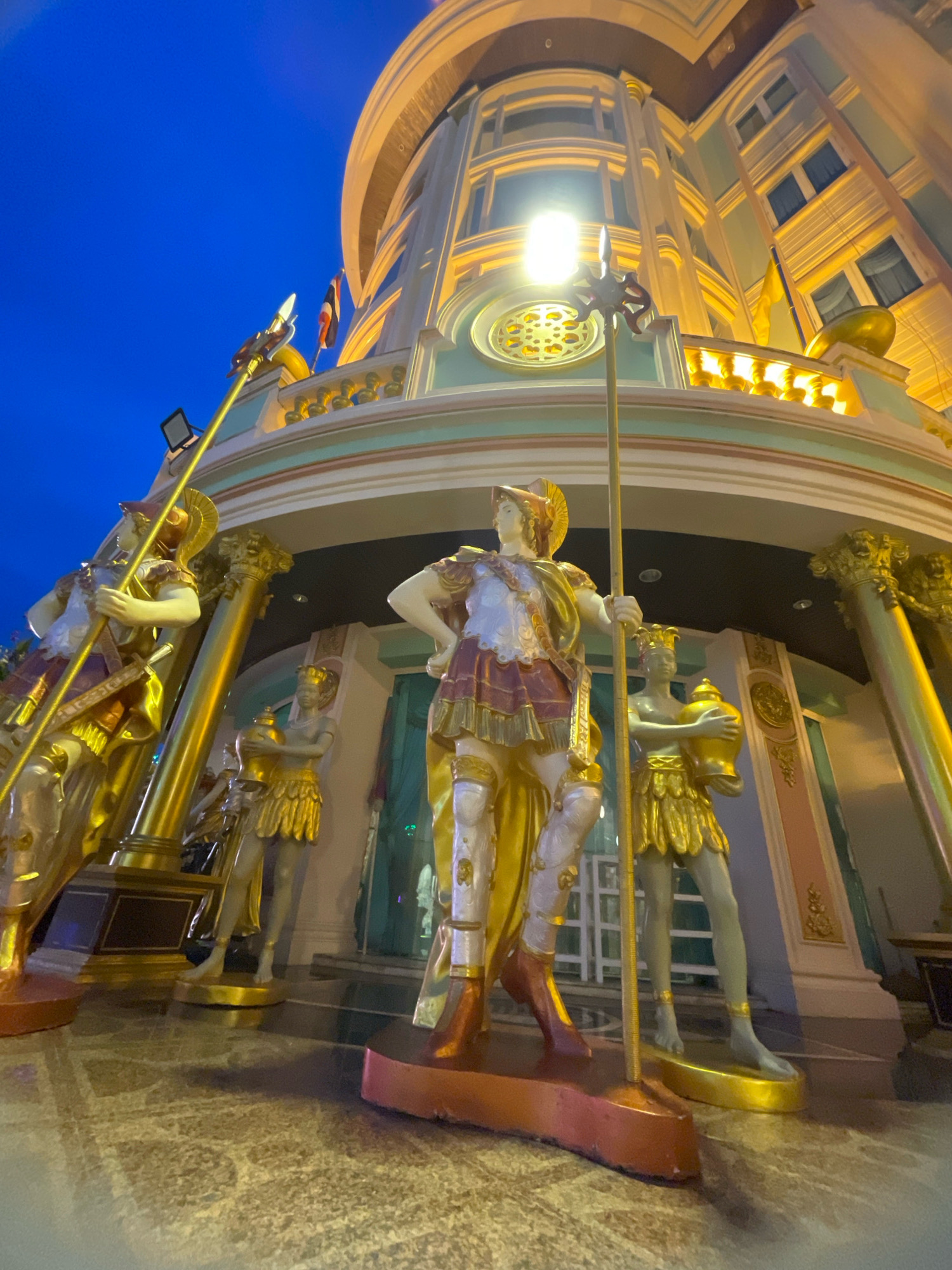 A low-angle, wide-shot photograph taken at dusk shows the exterior of a building in Pattaya City, Thailand, adorned with several life-sized golden statues of Roman-style figures. The statues are positioned in front of the building's entrance, with the central statue being the largest and most prominent. The building itself features a curved facade with multiple levels, ornate gold detailing, and windows that are illuminated from within. A bright light fixture is mounted on the building, casting a yellow glow. The sky is a deep blue, indicating the time of day is twilight or evening. The ground in front of the building is paved with a textured material. The overall impression is one of grandeur and opulence.