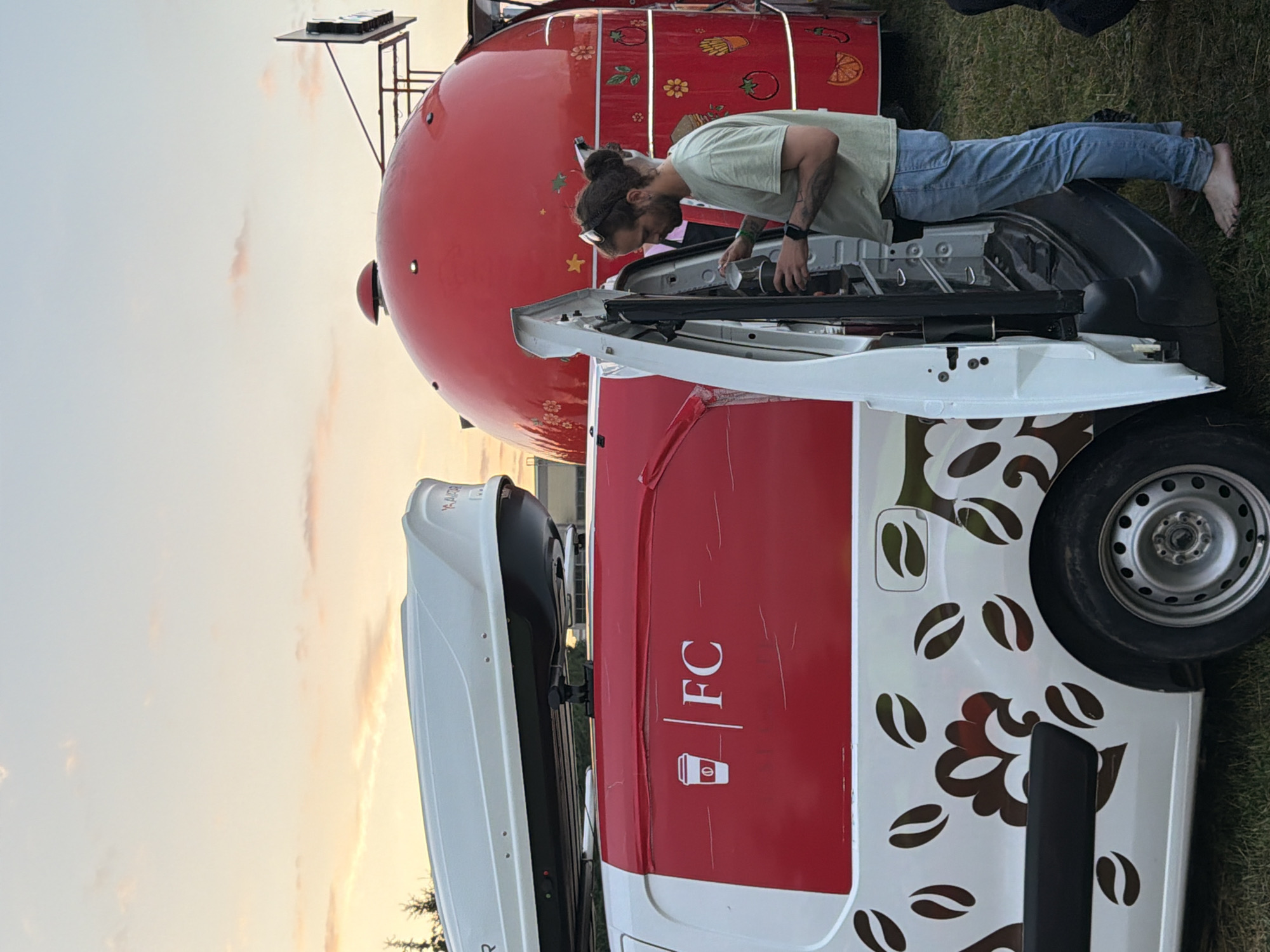 An exterior shot captures a man standing by an open van door, holding a cup and appearing to interact with the interior. The van, white with a red graphic band and coffee bean motifs, has a roof box. Behind it, a large, red, dome-shaped structure, possibly a food truck, is visible. The scene is set outdoors with a grassy ground and a sky with soft clouds. The lighting suggests it is either early morning or late evening.