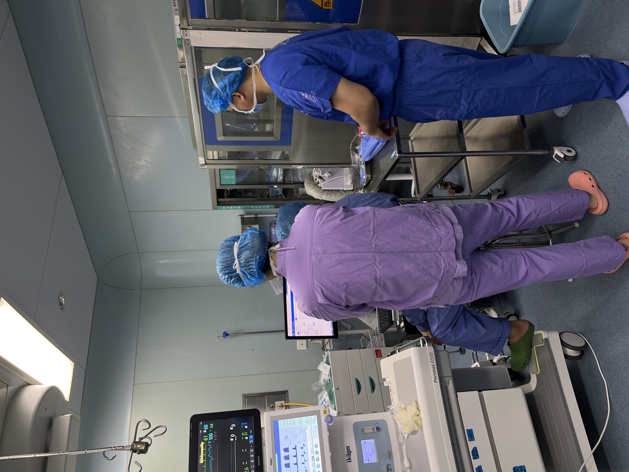 The image shows two medical personnel in an operating room or similar medical setting. The person on the left is wearing light purple scrubs and coral Crocs, while the person on the right is wearing blue scrubs and white Crocs. Both are wearing surgical caps. They appear to be attending to medical equipment, including monitors displaying vital signs and a Dräger Perseus A500 anesthesia machine. The environment is sterile and well-lit, with stainless steel surfaces and medical supplies visible. It is not possible to ascertain the exact location in Yuzhong District, China from the image.