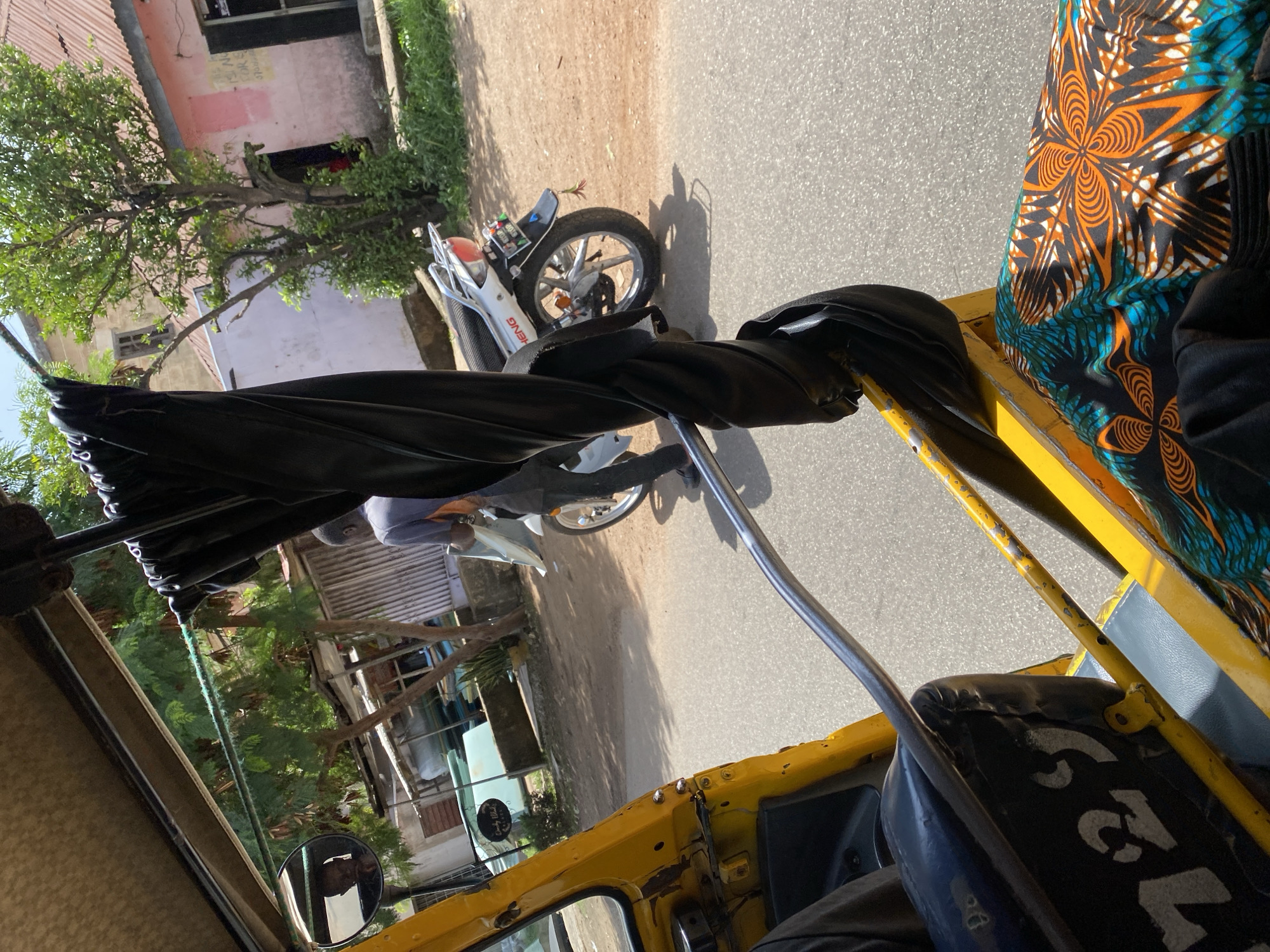 The image captures a street scene in Jos, Nigeria, from the perspective of a passenger inside a yellow auto-rickshaw. In the foreground, a black cloth is draped over a metal bar, partially obscuring the view. The passenger's legs, clad in vibrantly patterned African fabric, are visible. The auto-rickshaw's interior shows black seating with white numbering, possibly indicating a vehicle identification.
Outside the rickshaw, a person in a grey shirt and black pants is seen walking away from the camera, carrying a white object. They are standing next to a white motorcycle parked on a dirt and asphalt road. In the background, buildings with corrugated iron roofs and some greenery are visible. A circular sign with text is attached to a white structure. A side mirror reflects the face of the auto-rickshaw driver. The overall impression is a snapshot of daily life in a Nigerian city.