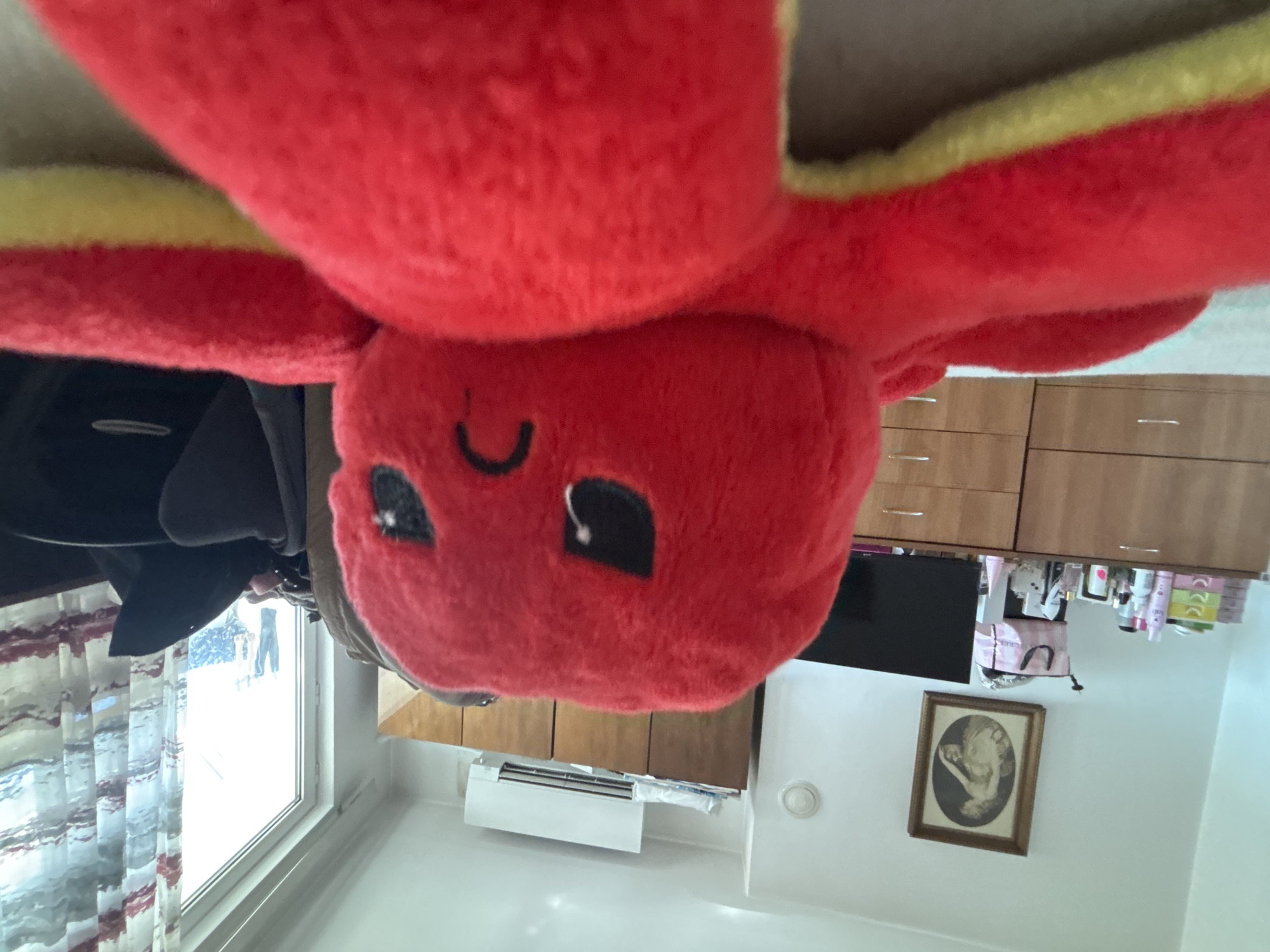 A close-up, slightly tilted image shows a red octopus plush toy with yellow trim in an indoor setting. The toy's face is visible with a curved, smile-like marking and two dark, square eyes.
In the background, a window reveals a snowy outdoor scene with indistinct figures. To the right, there are wooden cabinets and a TV screen, along with various cosmetic items. A framed portrait hangs on the white wall. An air conditioning unit is visible near the ceiling.
Given the presence of snow and the style of architecture visible through the window, the image appears to be taken indoors. Troyan, Bulgaria is not specifically identifiable from the provided information, but the general climate and architectural style align with such a location.