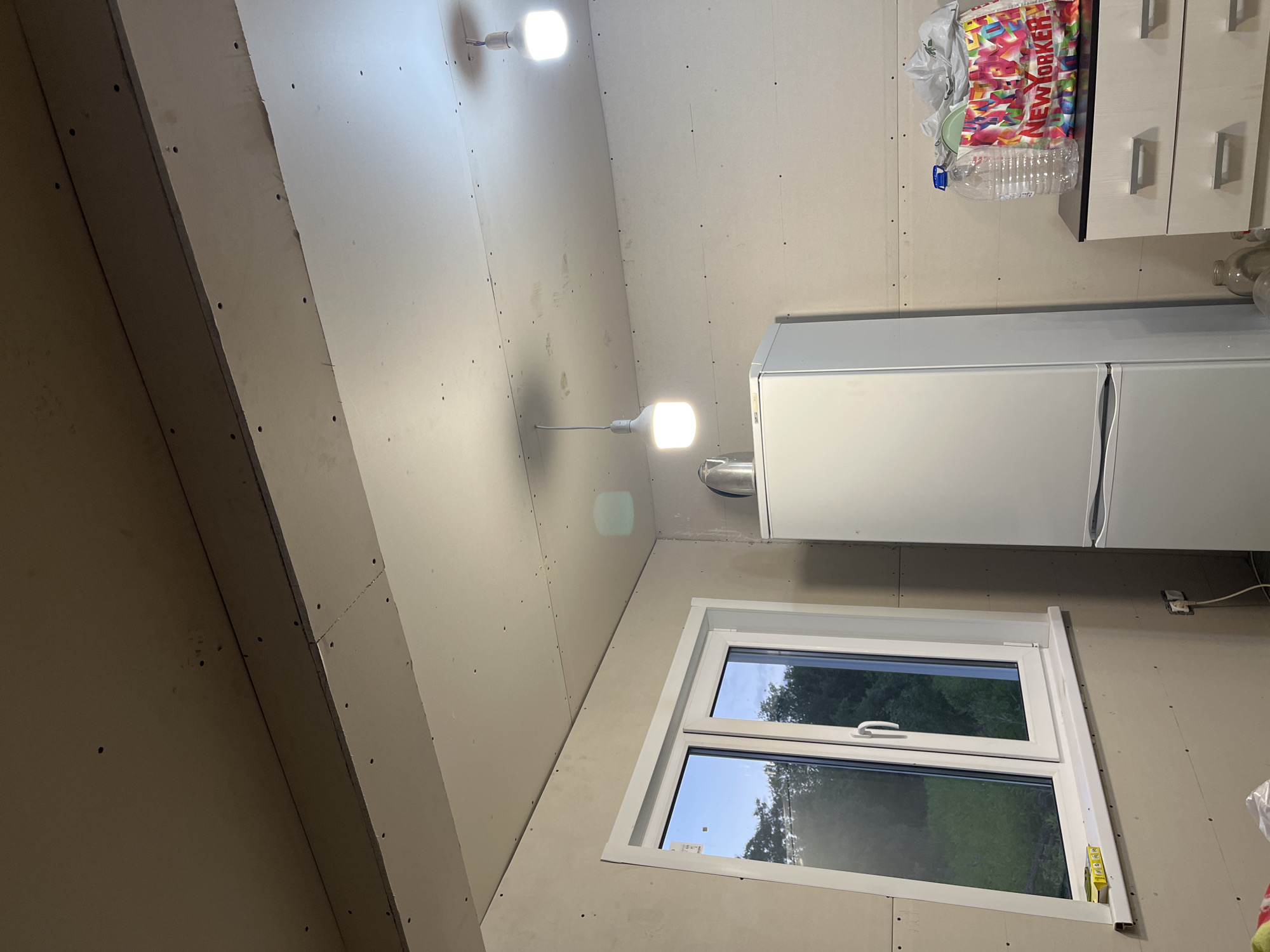 The image depicts an interior scene, likely a room under renovation, located in Клобушнево, Russia. The room is sparsely furnished, with an unfinished drywall ceiling and walls, evident from the exposed screw heads. Two modern pendant lights with white shades hang from the ceiling, illuminating the space. A white refrigerator stands against the right wall, and next to it, a black and white dresser with two drawers is visible, topped with a large clear plastic water bottle and a colorful tote bag from "New Yorker". A window with white framing is on the left side of the room, offering a view of green trees and the sky outside. The overall impression is one of a functional space in transition, possibly a kitchen or utility area. There are no people or animals in the image.