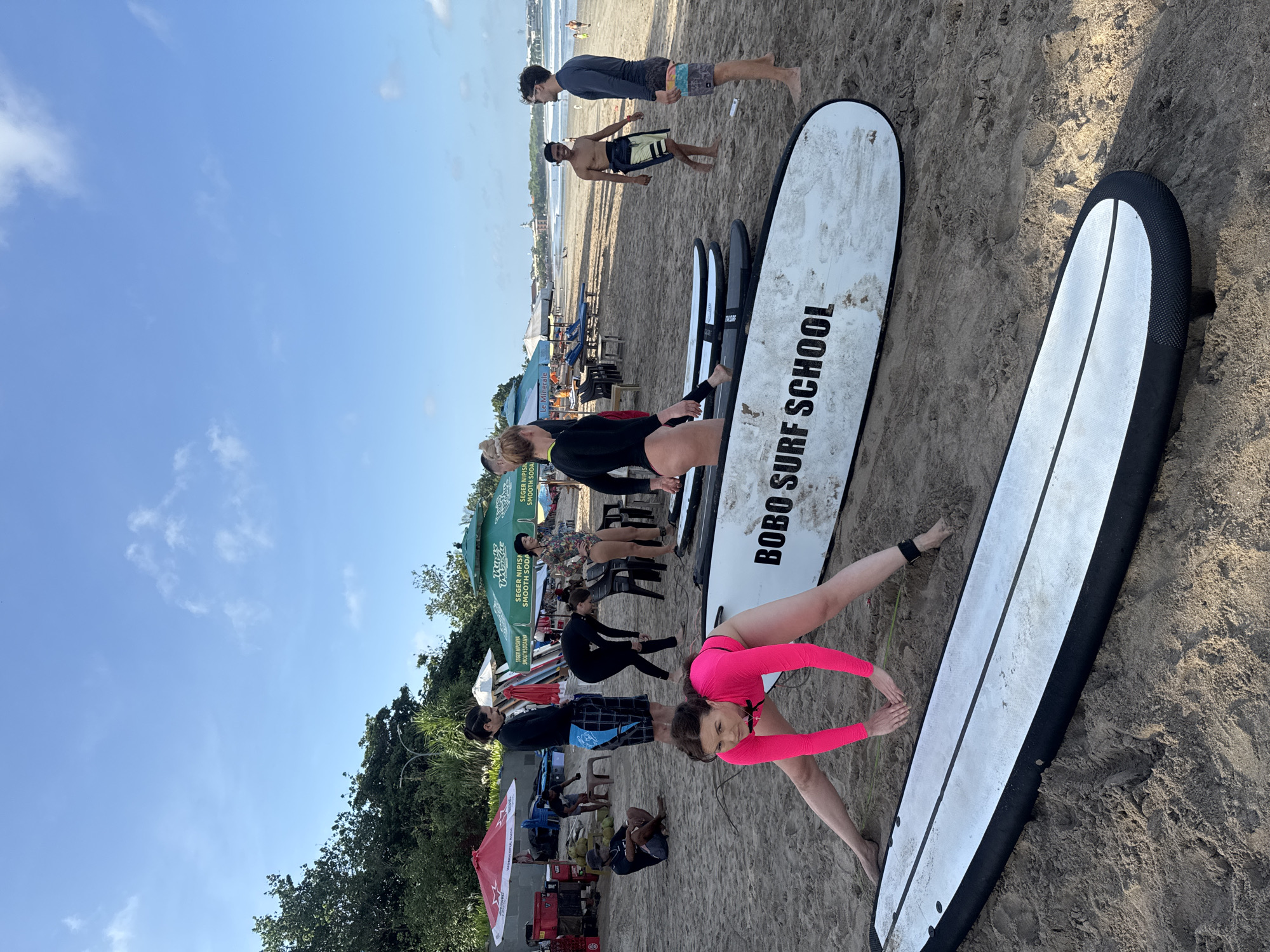 Here is a description of the image:
The image shows a beach scene, potentially in Kuta, Indonesia, as indicated by the "BOBO SURF SCHOOL" printed on a surfboard. Multiple surfboards are visible, some with people practicing or stretching on them. Several people are standing and sitting around. In the background, there are people on the beach, the ocean, and the sky. The scene includes beach umbrellas, tents, and vegetation.