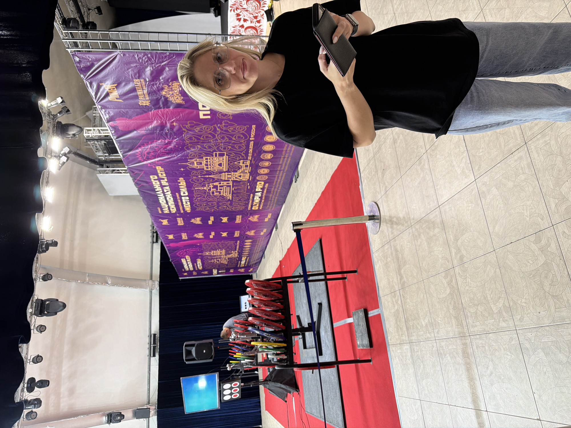 This media file captures a woman standing in front of a large purple banner advertising the "National Championship IPL/SPR 'Place of Strength' OLYMPIA PRO". The banner, written in Russian, also features imagery of buildings and text related to the event. The woman, wearing glasses, a black t-shirt, and blue jeans, holds a dark wallet or phone in her hand. The setting appears to be indoors, possibly a sports venue or exhibition hall, with a tiled floor and industrial lighting visible in the background. A red carpet leads to a display of weightlifting equipment, including red weight plates. The overall atmosphere suggests a sporting event is either about to commence, in progress, or has just concluded. The banner indicates the event took place on June 20-21, 2026, in Russia, specifically mentioning Suzdal.