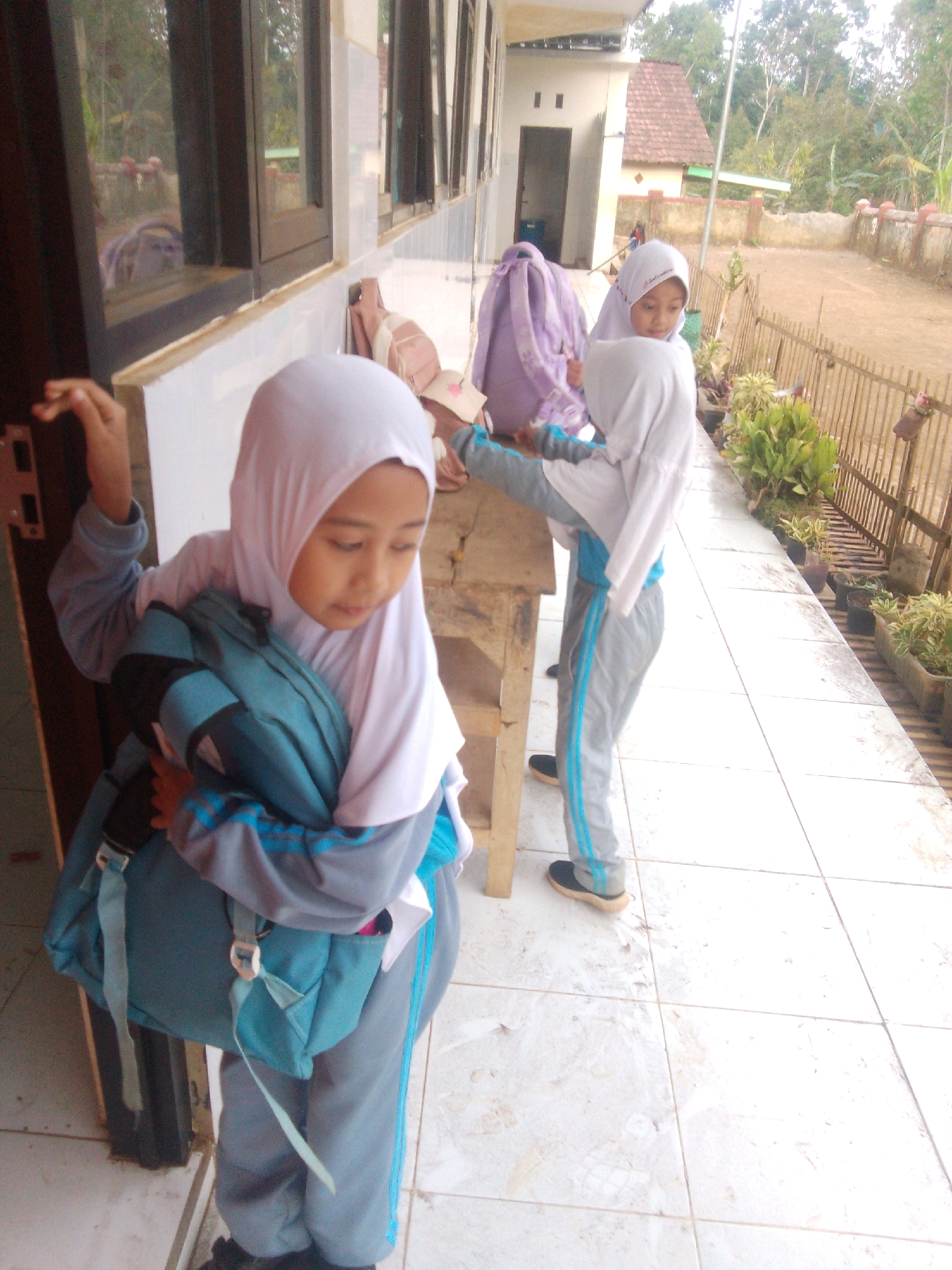 The image depicts a scene outside a school building, likely in Betek, Indonesia, given the surrounding vegetation and architectural style. Three young girls in school uniforms, which include headscarves, are present. The girl in the foreground is wearing a white headscarf and a grey and blue uniform, carrying a teal backpack. Her body is turned slightly away from the camera, and she appears to be interacting with something to her left, with her hand raised. To her right, another girl in a white headscarf and grey uniform is leaning against a wooden table, also with a teal backpack. A third girl, partially visible behind the second, wears a similar uniform and a purple backpack. The background shows a dirt yard, a wooden fence with potted plants, and the school building with windows. The ground is tiled. The lighting suggests it is daytime.