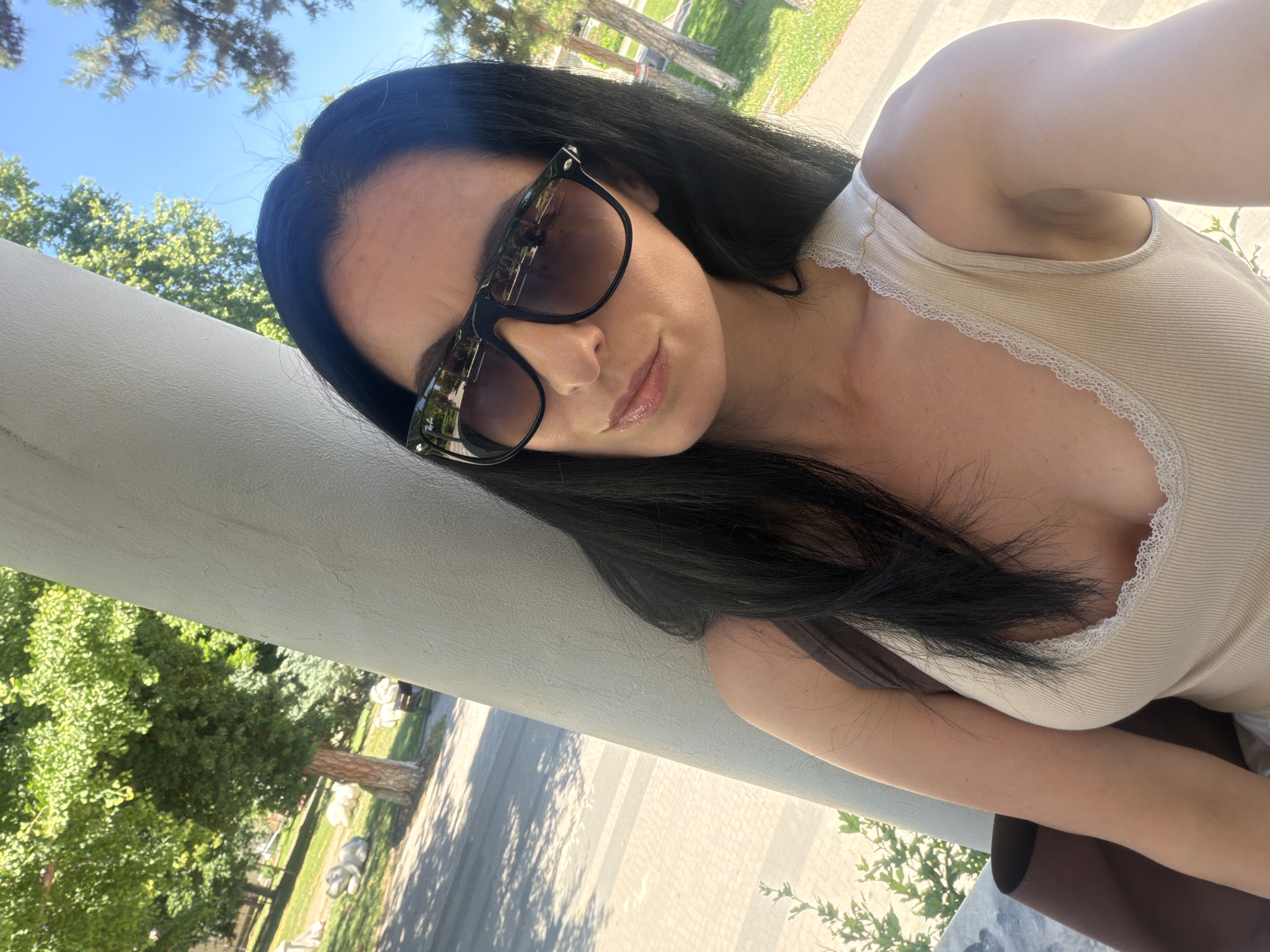A woman with long, dark hair and sunglasses is taking a selfie outdoors. She is wearing a light-colored tank top with lace trim. The background shows trees, grass, and a paved area, suggesting a park or outdoor public space. The specific location in the city of Pleven, Bulgaria, is not identifiable from the visual information provided.