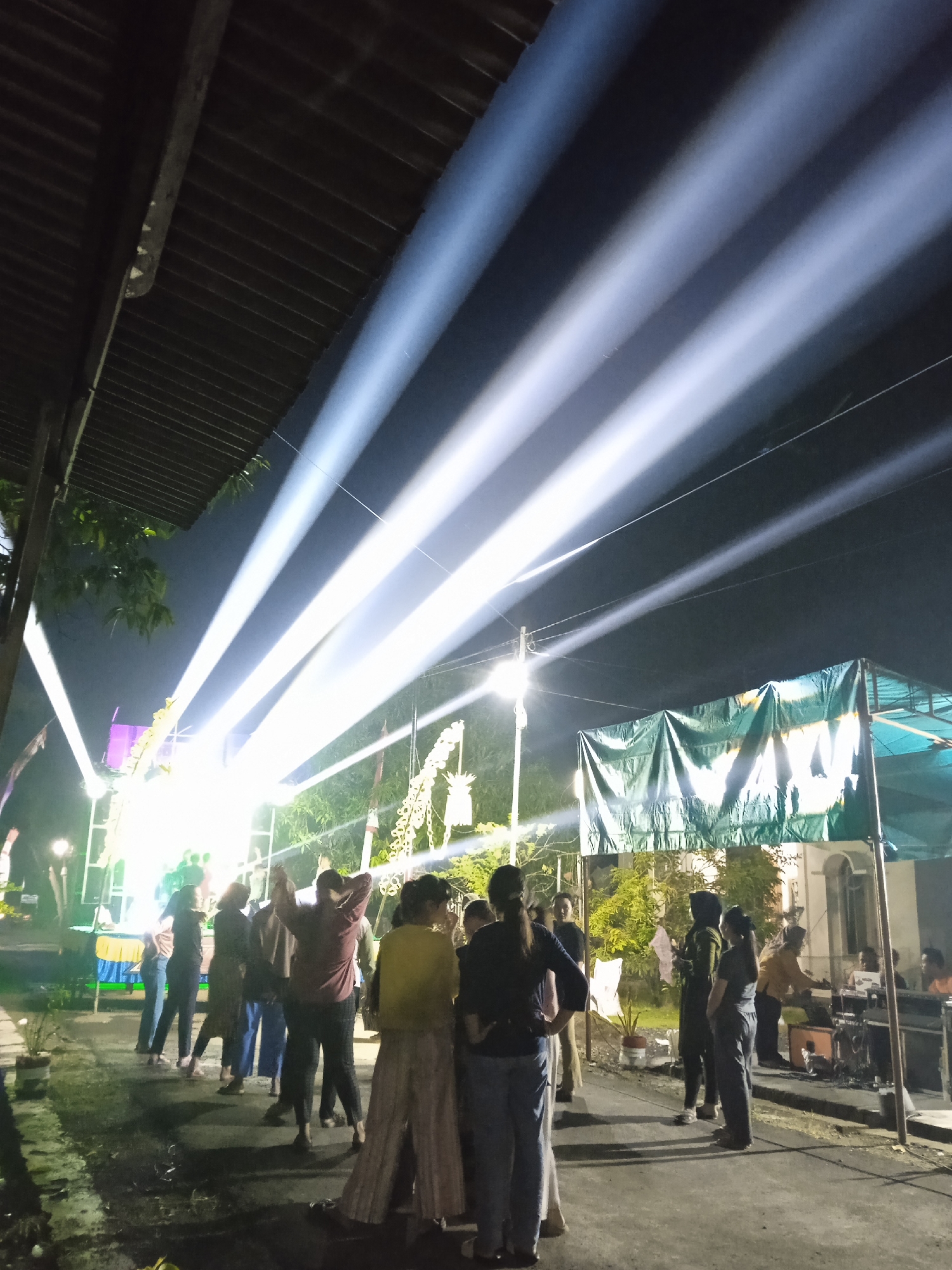 The image depicts a nighttime outdoor event with people gathered near a stage and a tent-like structure. Intense white light beams shoot upwards into the dark sky. People are standing and interacting in groups, some appearing to face the stage. A structure with a corrugated roof is visible on the left side of the frame. The event is occurring in Winong, Indonesia.