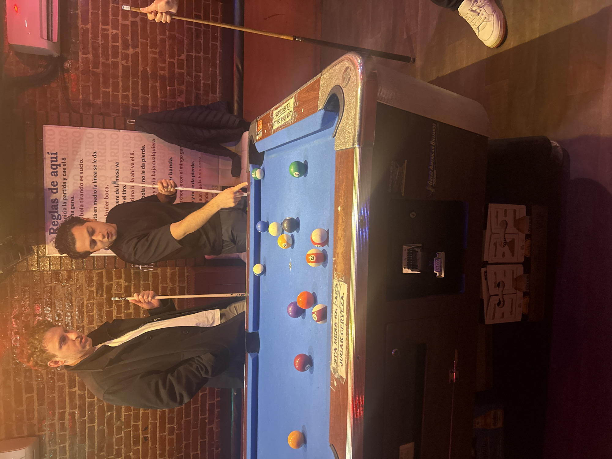 The image shows two people playing pool in a dimly lit bar. A pool table dominates the center of the frame, with several billiard balls scattered across its blue surface. Two men are positioned around the table, one appearing to be lining up a shot with a pool cue, while the other watches. The bar has exposed brick walls. A sign with pool rules is visible on the wall behind the players. A pool table mechanism shows a $2.00 charge for the table. The bar may be located in New York City, United States of America.