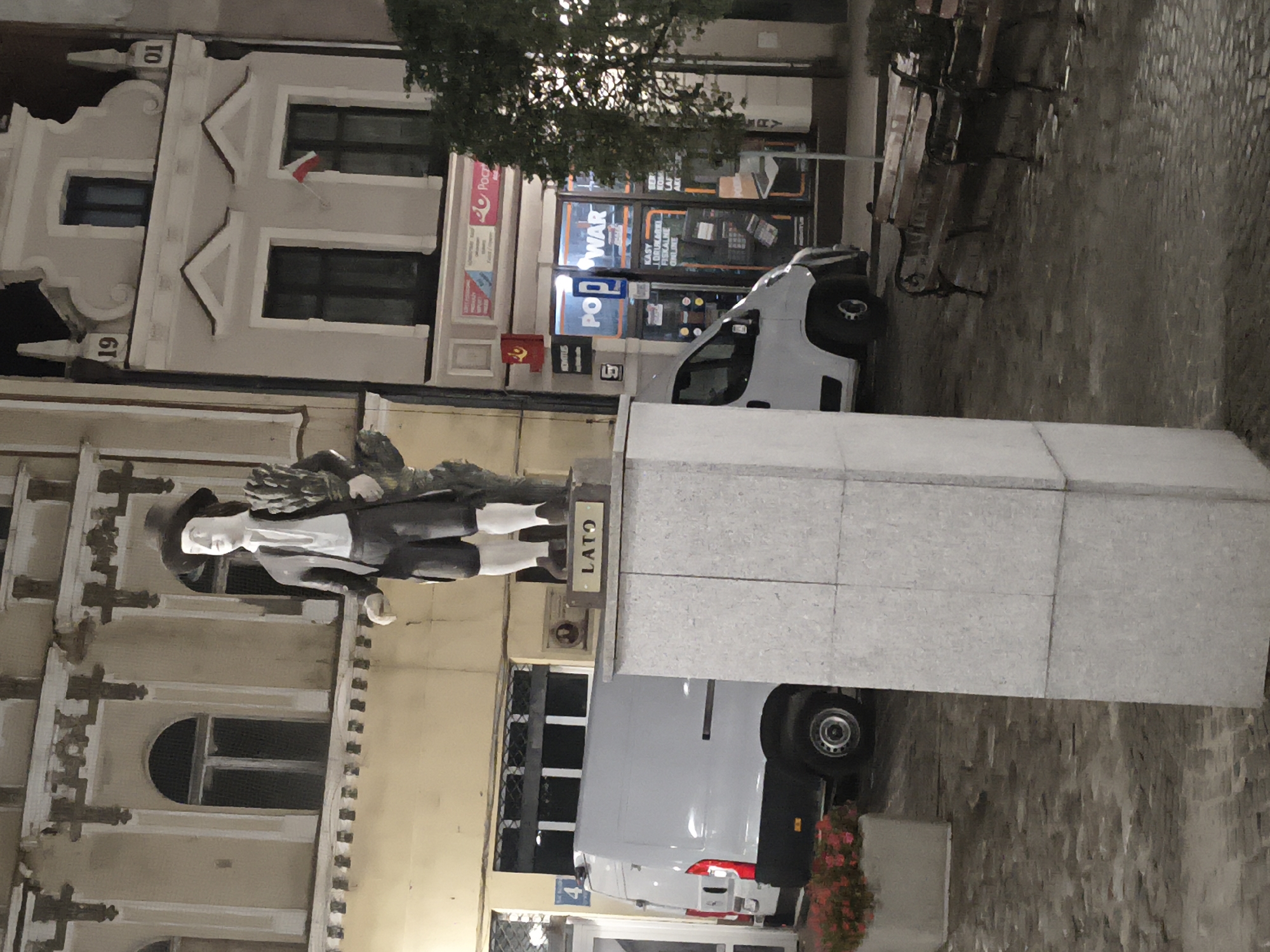 A statue of a man labeled "LATO" stands on a pedestal in front of a light-colored building. The statue depicts a man wearing a hat, white shirt, and dark shorts, holding a bundle of wheat. To the left of the statue, a white van is parked. The building behind the statue has arched windows and decorative architectural elements. The street in front of the building is paved with cobblestones. In the background, other buildings are visible, including one with a sign reading "POZ WAR". Benches are situated on the right side of the image. The scene appears to be at night or in low light conditions. This statue is located in Tczew, Poland, specifically on Długa Street, honoring Stanisław Lato, an important figure in the history of Tczew.