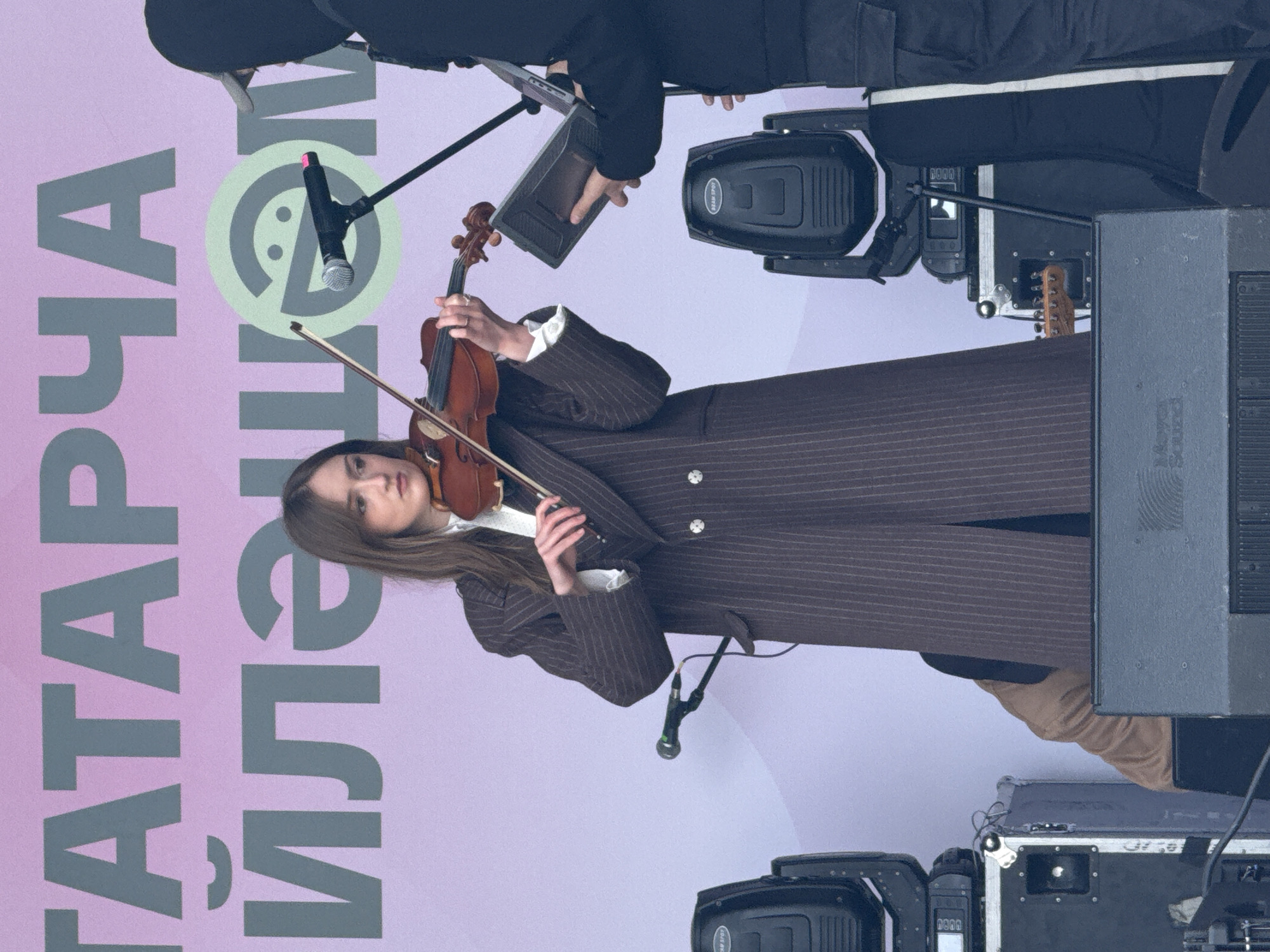 A female violinist plays on an outdoor stage in Kazan, Russia. She wears a pinstripe coat. A microphone stands before her. Behind her is a backdrop with the words "ТАТАРЧА ИЛҺАМ" in Cyrillic script. Technical stage equipment, including lighting fixtures and speakers, surrounds the performer. Another person is visible above the violinist, potentially adjusting equipment.