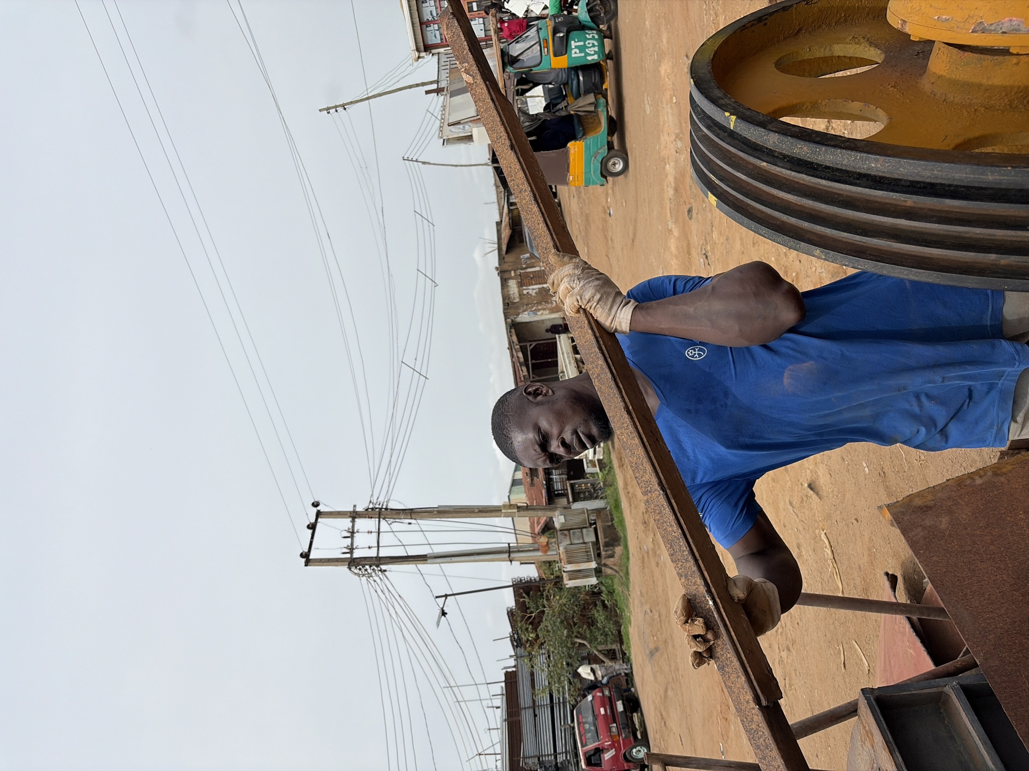 A man in a blue shirt and gloves is holding a long metal beam in an outdoor environment in Gura Top, Nigeria. Behind him, power lines and poles are visible against a cloudy sky. A yellow wheel and a red truck are also in the scene. A green and yellow public transit vehicle with "PT 1495" on it is visible as well. The ground is dirt.