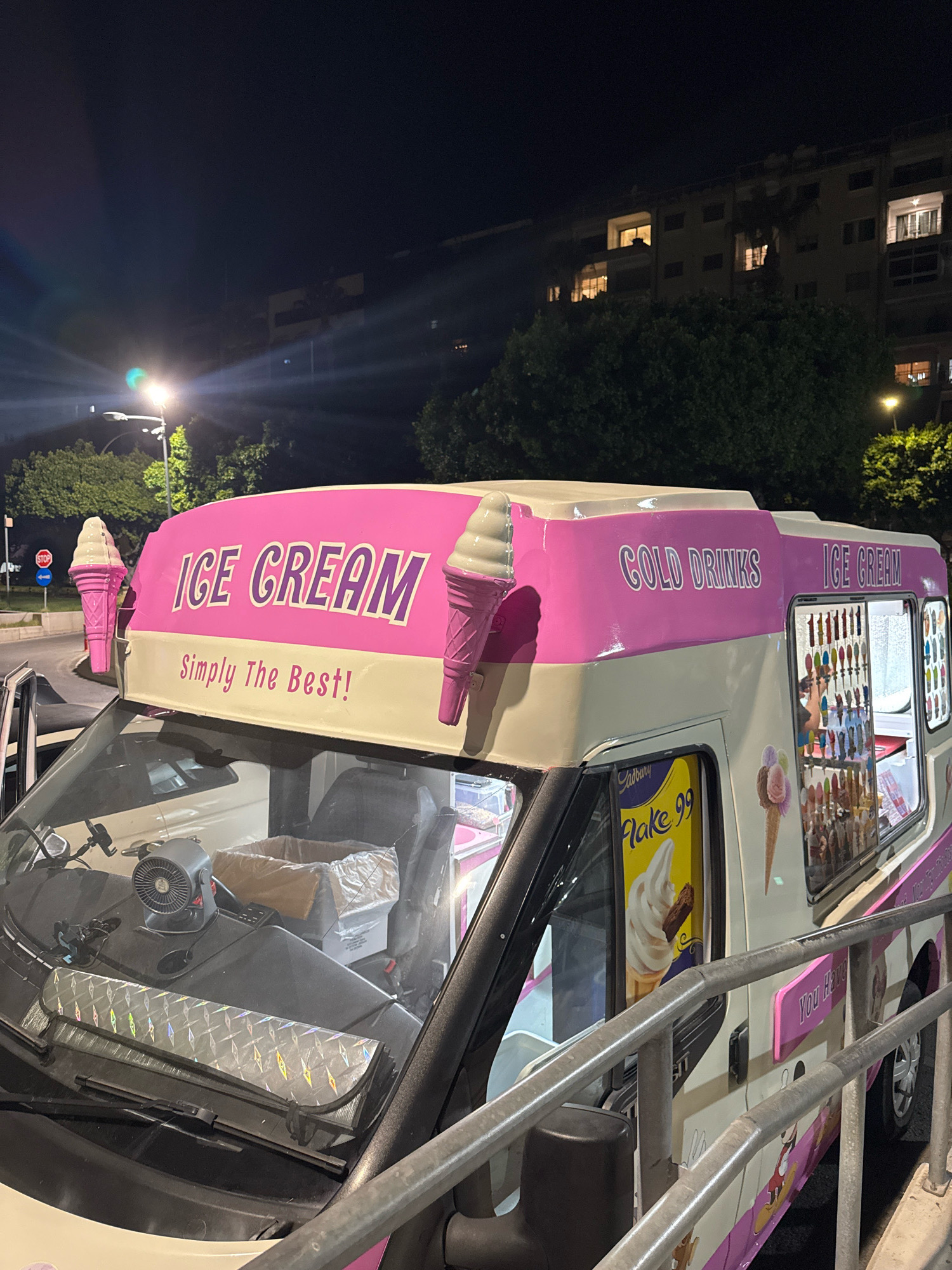 A white and pink ice cream truck is parked at night on a street in Limassol, Cyprus. The truck has "ICE CREAM" and "COLD DRINKS" written on its side, as well as an image of a Cadbury Flake 99. In the background, there is a multi-story apartment building with lit windows and trees. The street is illuminated by streetlights.