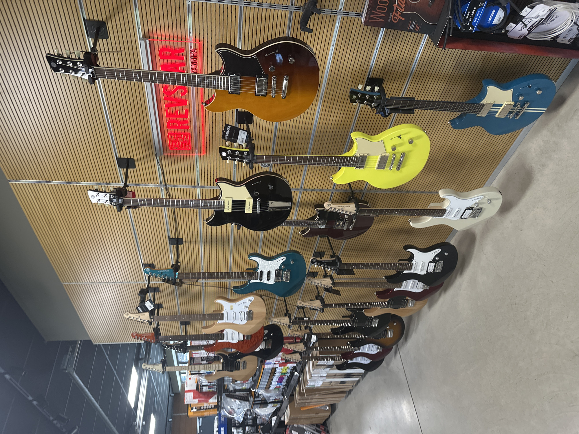 The image shows a wall display of electric guitars in a music store. The guitars are of various colors and styles, mounted on black wall hooks against a light brown slatted wooden background. In the upper right corner, a red neon sign that reads "REVSAR YAMAHA" is visible. Below the guitars, shelves are stocked with various musical accessories. The floor is a light gray concrete. The scene appears to be an indoor retail environment, likely a guitar shop, with no people visible. The context of Sofia, Bulgaria is not evident from the image itself.