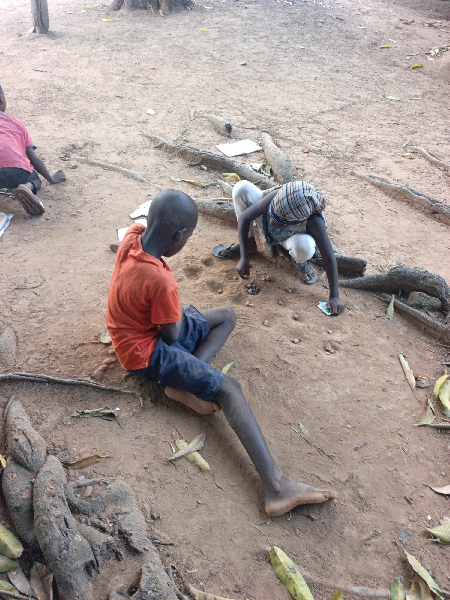 The image depicts three children in an outdoor setting with bare earth ground, tree roots, and scattered leaves. One child sits in the foreground, wearing an orange t-shirt and blue shorts, with their back to the camera. Another child, wearing a patterned head covering, white pants and sandals, bends over, seemingly examining the ground. A third child is partially visible in the background, wearing a red shirt. Paper and debris are scattered on the ground. The location is Akwanga, Nigeria.