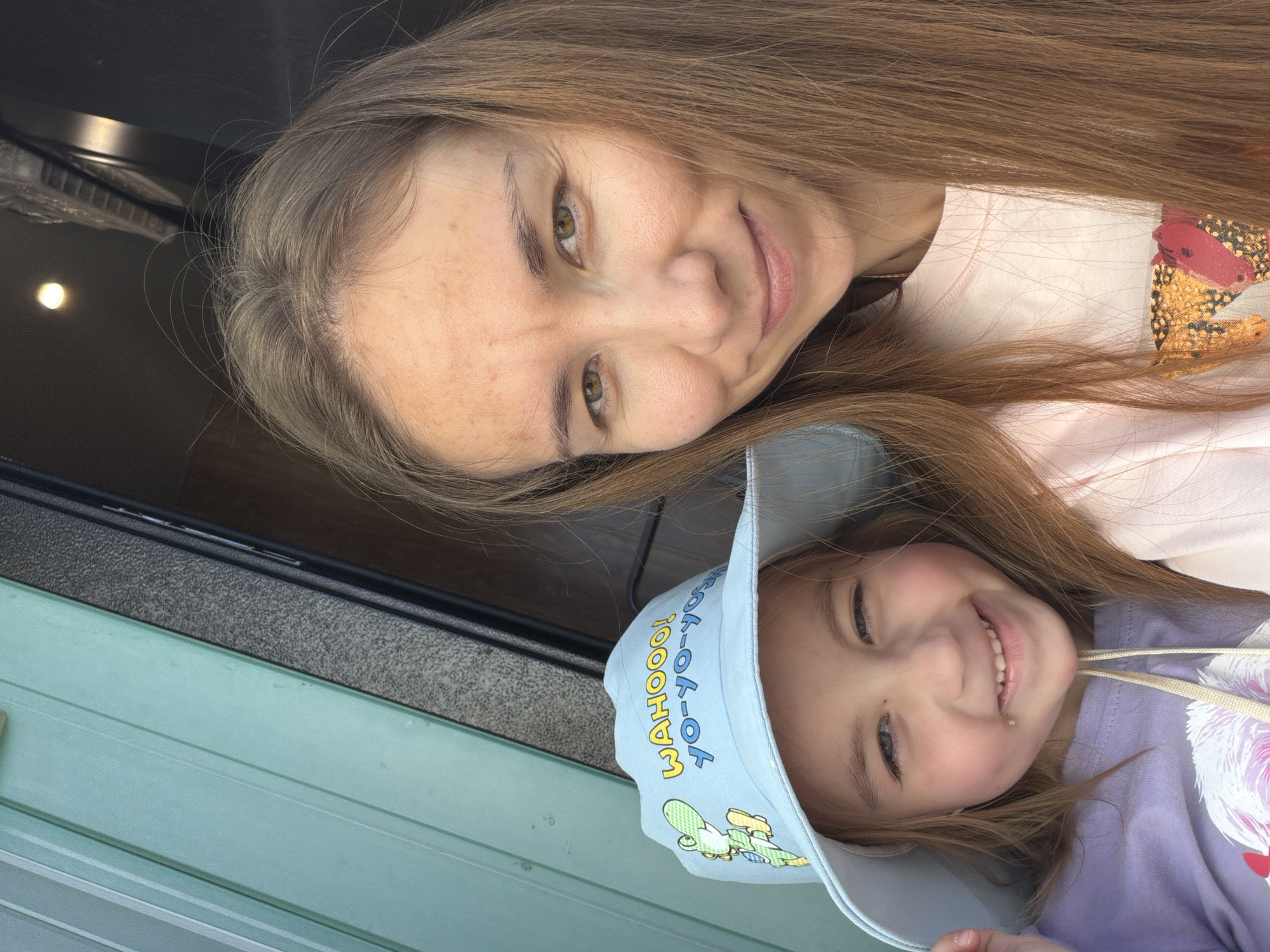 A medium shot captures a mother and daughter posing for a selfie. The daughter, wearing a light blue bucket hat with a cartoon character and text "WAHOOO! YO-YO-YO!", smiles widely and leans into the frame from the left. She is wearing a lavender t-shirt. The mother is positioned to the right of the daughter, her face close to the camera, and she is also smiling. She has long, light brown hair that frames her face. Both are wearing casual clothing. The background appears to be outdoors, with a light blue, ribbed surface on the left and a darker, textured surface on the right, suggesting an outdoor structure or wall. The lighting suggests it is daytime. The image does not contain enough information to determine the specific location in Tambov, Russia.